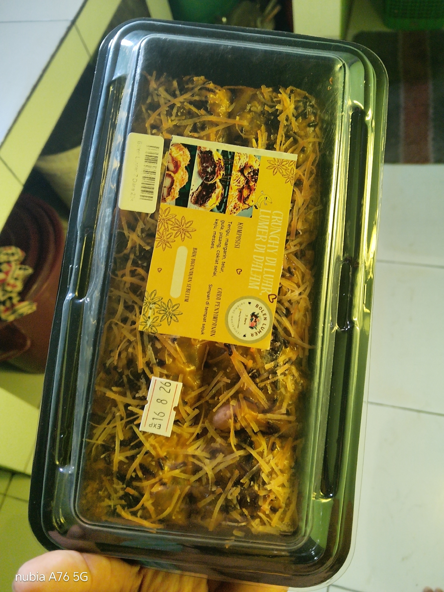 The image shows a clear plastic container filled with a pastry item. The pastry appears to be topped with shredded cheese and dark-colored filling, possibly chocolate. A yellow label with text and images of pastries is affixed to the container. Another smaller label with a barcode and text is also visible. A "EXP 16 8 26" sticker is affixed to the container. The container is placed on a tiled surface. The background is out of focus and shows what appears to be a floor and a wall. The location is identified as Cakung, Indonesia, based on the context provided.