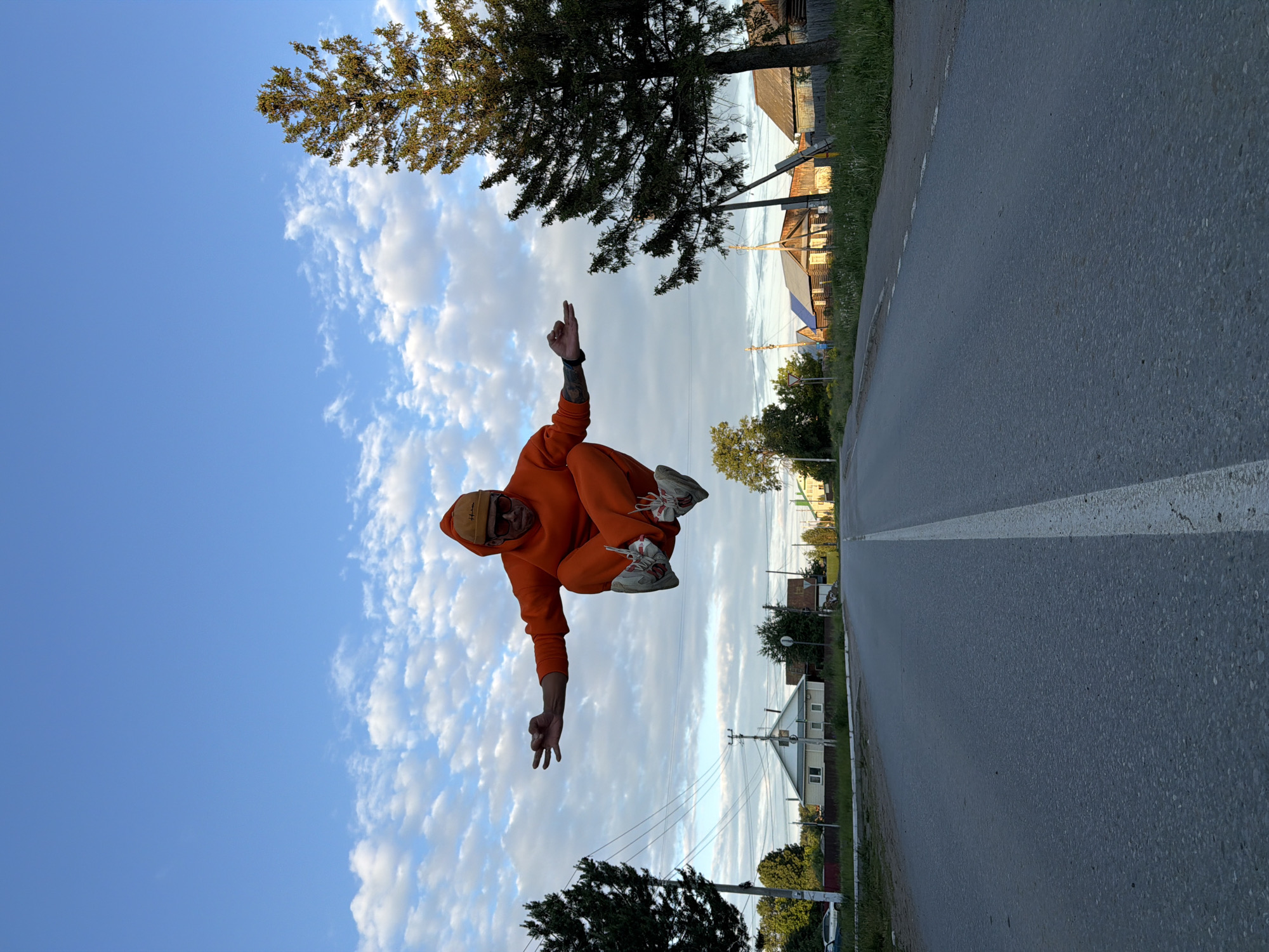 The original image, rotated 90 degrees clockwise, captures a person mid-air, seemingly in a jump or float, against a semi-rural backdrop in Chaikovsky, Russia. The central figure is dressed in a vibrant orange matching hoodie and sweatpants, light-colored sneakers with red details, and a light brown baseball cap that clearly displays the word "Hooligan." Tattoos are visible on their left forearm. The person is performing a hand gesture with two fingers raised on their right hand and an "OK" sign with their left hand, conveying a sense of playfulness or confidence.
The scene unfolds along an asphalt road marked with a white dashed line. Grassy shoulders flank the road, leading to a landscape dotted with various trees—some evergreen, others with deciduous foliage. Several houses, featuring wooden construction and traditional roofs, are visible in the background, interspersed among the trees. Utility poles and overhead wires further indicate a settled area. The sky above is partly cloudy, showcasing patches of clear blue, suggesting it is daytime, possibly late afternoon or early evening given the soft, ambient light. The image captures a dynamic human element within a peaceful, transitional setting between nature and human settlement.