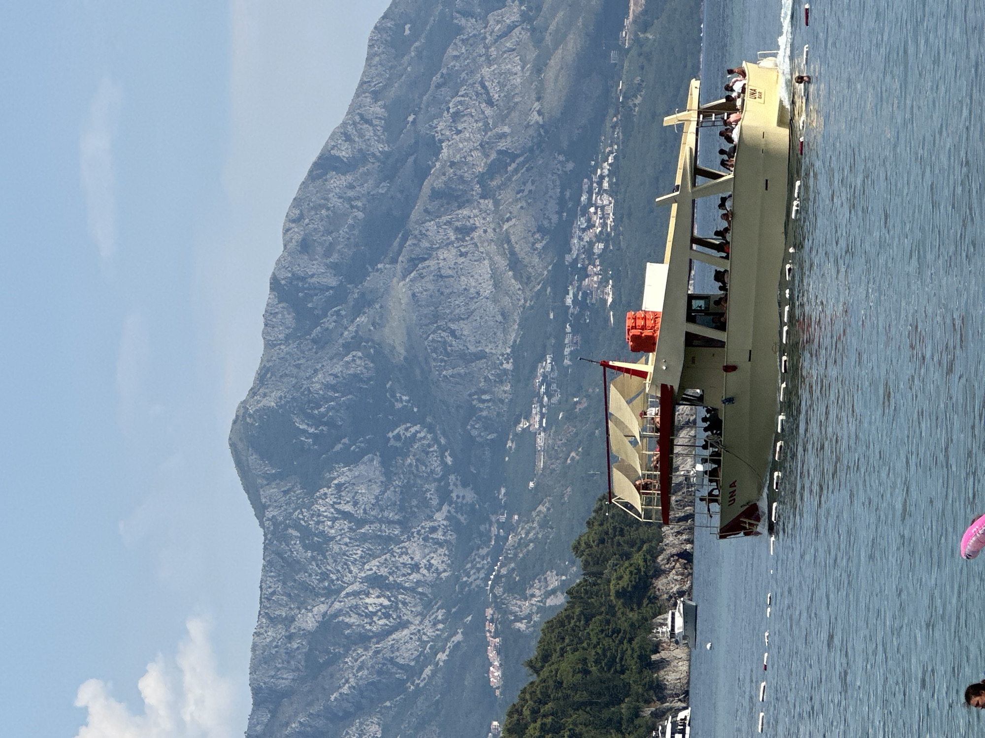 The image depicts a scenic view of a boat on the water with a mountainous backdrop. The boat, labeled "UNA BAR," is light-colored and appears to be carrying passengers. Several people are visible on the deck, enjoying the ride. The boat has multiple levels, with some passengers seated under awnings. A red life preserver is visible on the side of the boat.
In the background, a large, rugged mountain dominates the scene. The mountain is covered in vegetation on its lower slopes, and a small town with white buildings is scattered along the mountainside. The water is a calm, blue color, with gentle ripples. A few white buoys are floating in a line on the water. A pink inflatable object is partially visible in the lower right corner. The sky is clear and blue, with a few wispy clouds. The overall impression is a sunny day in a coastal location. The image is taken from a slightly elevated perspective, looking towards the boat and the mountain. The orientation of the image is portrait, with the boat running vertically. The scene evokes a sense of leisure and tourism. The time of day appears to be daytime, likely midday, given the brightness of the sunlight. The location context provided is Budva, Montenegro, which aligns with the mountainous coastal scenery.