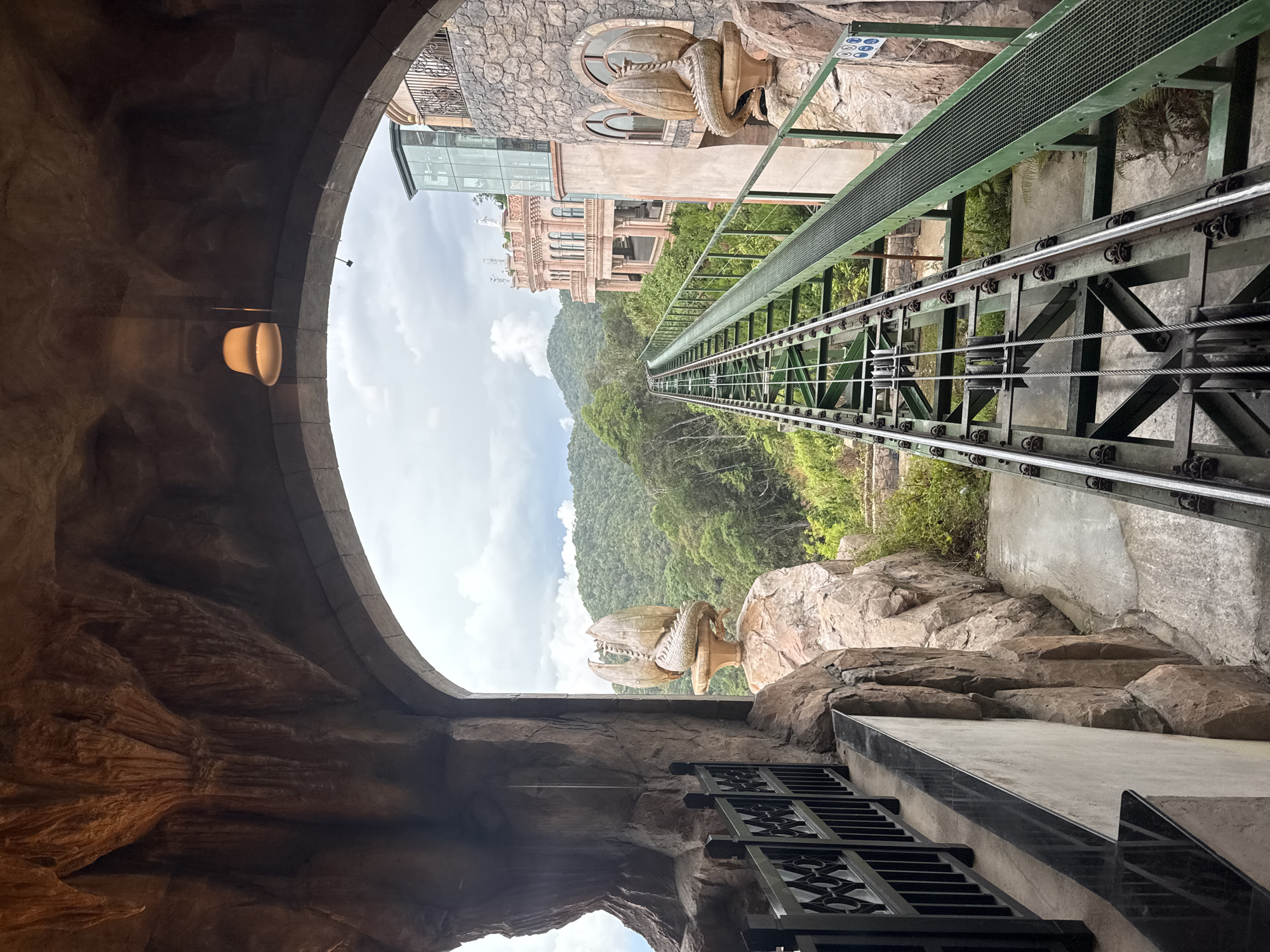 The image depicts a steep cog railway track leading downhill through a mountainous, forested landscape. The track is flanked by rocky terrain with built structures, including a building complex on the left and a dragon statue on both sides, visible through an archway/tunnel-like structure. The architecture appears to be fantastical, medieval-themed. The sky is cloudy. The cog railway is located in Ba Na Hills, Đà Nẵng, Vietnam.