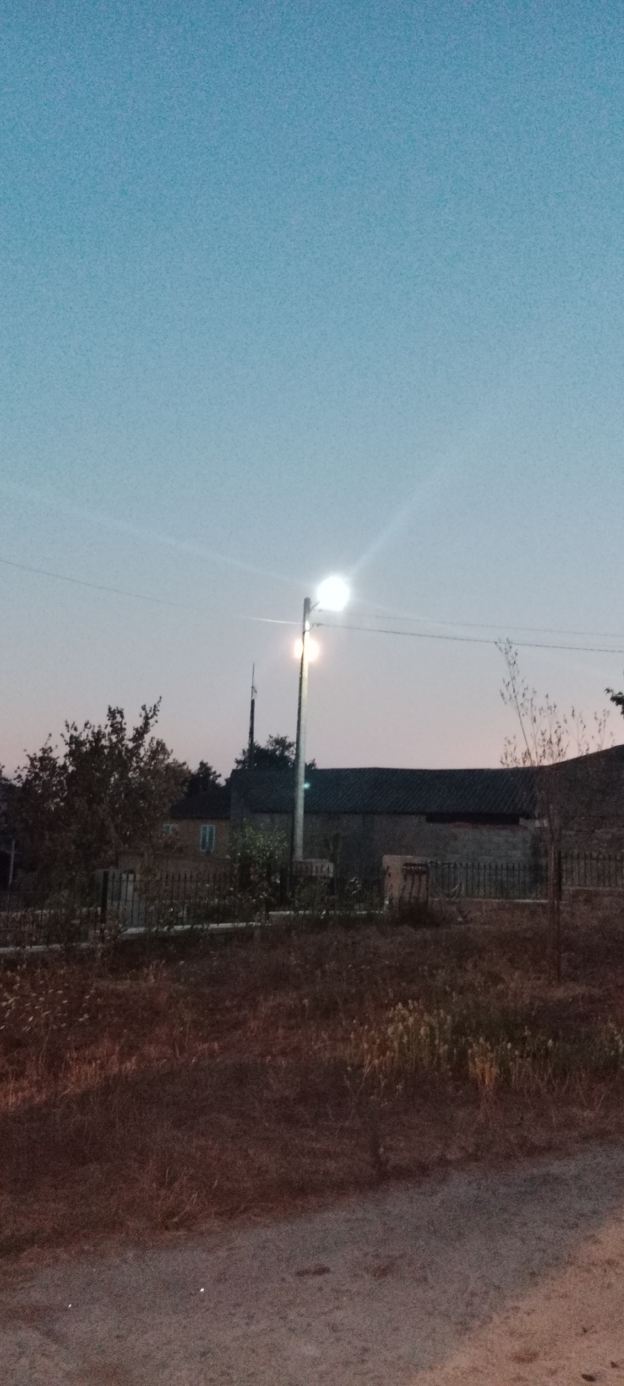 The image depicts a dimly lit outdoor scene, possibly at dusk or dawn, in Adai, Spain. The sky is a gradient of soft blue and light pink. A single streetlight with a bright, glowing bulb dominates the mid-ground, casting a sharp light. Power lines extend horizontally across the frame, intersecting the light pole. Below the light, a row of buildings with dark roofs and subtle window details are visible, separated from the foreground by a low, dark fence. To the left, a cluster of trees with dense foliage stands out. The foreground consists of dry, brown grass and dirt, with some scattered white wildflowers. The overall atmosphere is calm and still, with no people or animals present.