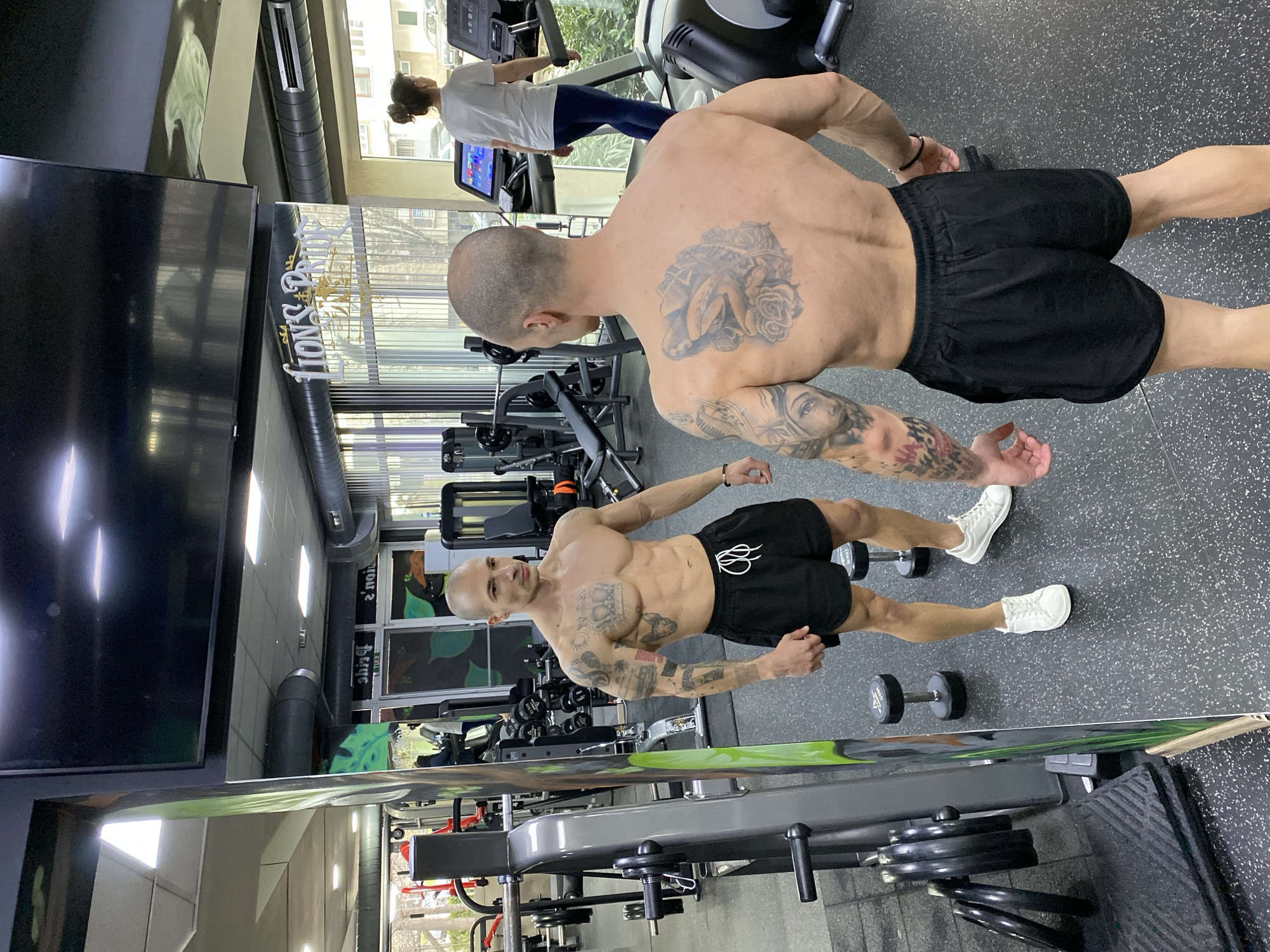 The scene depicts the interior of the Lion's Pride Fitness & Spa gym, located at Ulitsa "Gorni Dybnik" 10, Pleven, Bulgaria. A bald, muscular male subject stands shirtless in the foreground, facing his reflection in a large, floor-to-ceiling mirror. He wears black athletic shorts with white drawstrings and white athletic shoes. His back is toward the viewer, revealing a large tattoo depicting a figure amidst roses on his upper back, and extensive sleeve tattoos on both arms. His reflection in the mirror displays his front, revealing a toned physique with visible abdominal muscles and additional chest tattoos, including a crown and other various designs. In the background, to the right, a female subject with her hair in a bun walks on a treadmill; she is wearing a white t-shirt and dark blue leggings. The gym features dark speckled rubber flooring, various weight machines, dumbbells (including a 12 kg dumbbell near the male subject's feet), and weight racks. Large windows with vertical blinds line a far wall, admitting natural light. The ceiling contains recessed lighting and exposed dark ventilation ducts. A large black television screen is mounted above the mirror. The gym's name, "LION'S PRIDE," is visible in stylized gold lettering as a reflection on the mirror surface.