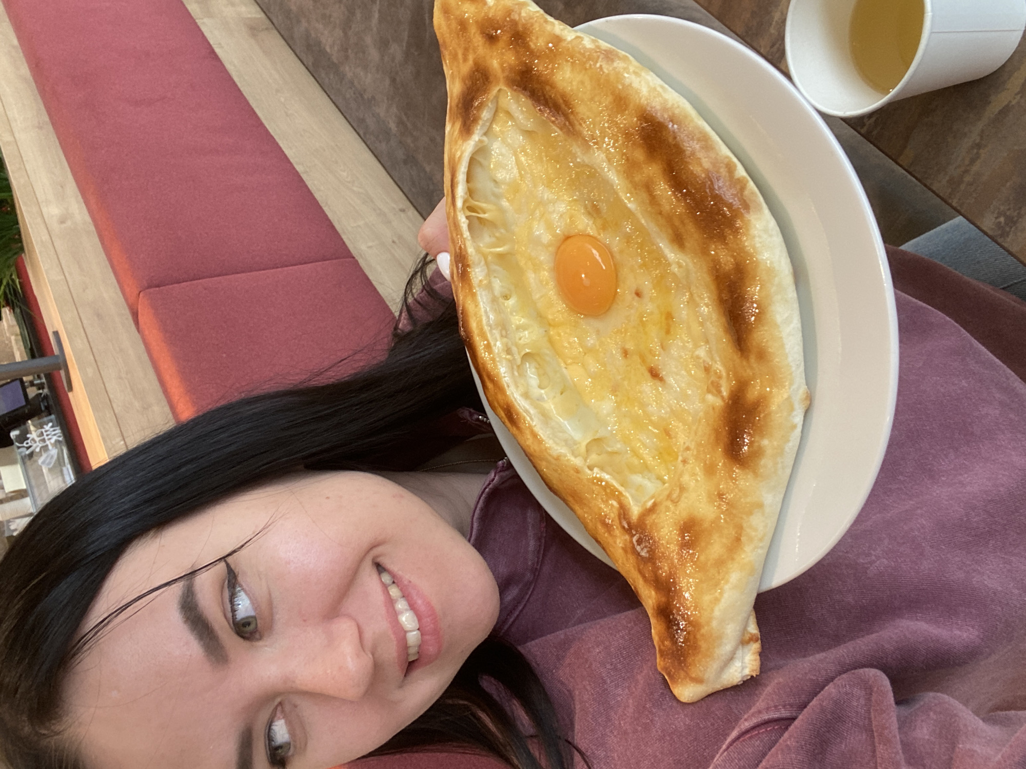 A selfie of a young woman with black hair smiling, likely indoors, with a Khachapuri Adjaruli on a white plate positioned in front of her. The khachapuri has a baked crust, melted cheese, and a raw egg yolk in the center. Next to the plate is a white cup containing a yellow liquid. The woman is wearing a dark pink or purple shirt. The background shows a bench with a red cushion and a wooden surface. The location is identified as Perm, Russia based on the Khachapuri Adjaruli.