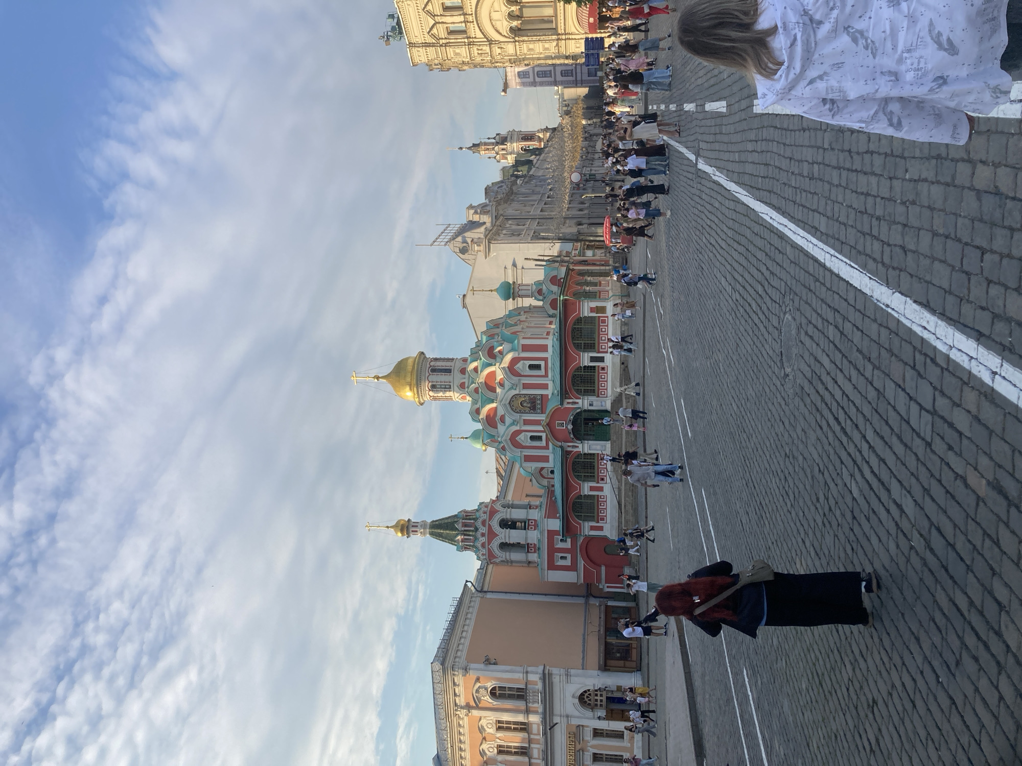 This image depicts a view of Red Square in Moscow, Russia, with the colorful Saint Basil's Cathedral prominently featured in the center. The cathedral boasts a distinctive onion-domed roof, a green and red facade, and intricate architectural details. Surrounding the cathedral are other historical buildings, including structures with classical architecture and golden domes, typical of Russian Orthodox churches. A cobblestone pavement covers the ground, indicating the square's historical significance. Numerous people are seen walking and gathering in the square, engaged in various activities such as sightseeing and taking photographs. The sky above is partly cloudy, with soft sunlight illuminating the scene, suggesting it is either early morning or late afternoon. The overall atmosphere appears lively and public, characteristic of a major tourist attraction.