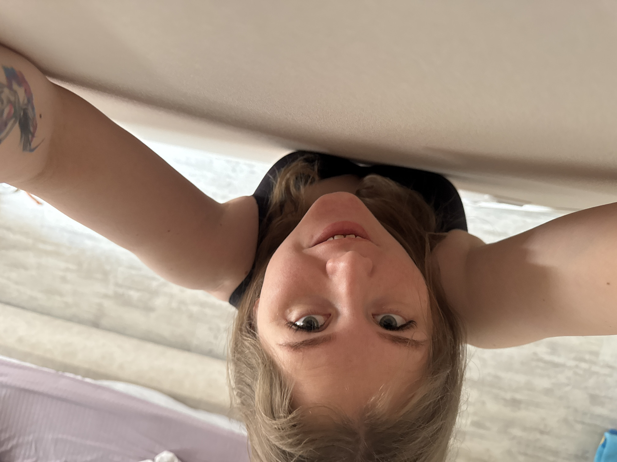 A selfie-style photograph taken from a low, upward angle features a light-skinned person with blonde hair. The individual's face, partially illuminated, is in focus, revealing green eyes, a slightly open mouth, and a view up their nostrils. The person appears to be wearing a black tank top. The background includes a white ceiling and a light-colored wood-like floor. A portion of a purple bed or sofa is visible in the lower left corner. A tattoo is visible on the person's left arm. The photograph appears to be taken indoors. The location details in the city of Gornyi (Dvurechenskoe S/P), Russia are not discernible from the image itself.