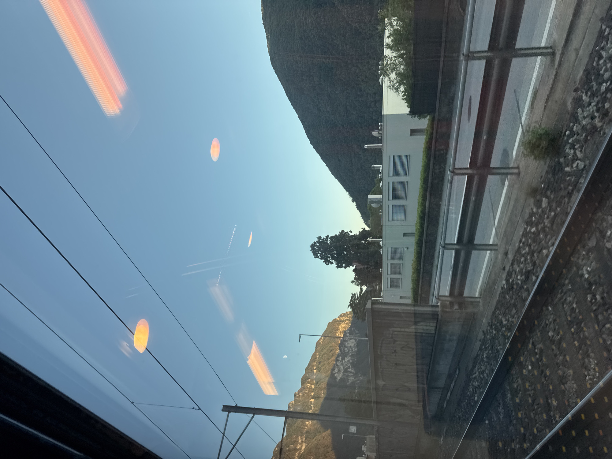 The image captures a view from a moving train window, looking out towards a scenic landscape. The scene depicts train tracks in the foreground, with ballast and a section of the train's interior visible at the bottom edge. Beyond the tracks, there is a road with a guardrail, followed by a modern, light-colored building with louvred windows and what appear to be ventilation units on the roof.
The backdrop consists of two distinct hillsides. The left hillside is steep and rocky, partially illuminated by sunlight, casting shadows. A faint crescent moon is visible in the sky above this hillside. The right hillside is covered in dense vegetation.
The sky is a clear, pale blue, with reflections of orange and yellow light streaks visible on the train window, suggesting the presence of external light sources or reflections of the train's interior. Two circular orange-yellow light reflections are prominent in the upper portion of the sky. The overall impression is one of a serene, daytime journey through a mountainous region. The location is identified as Val Mara, Switzerland.