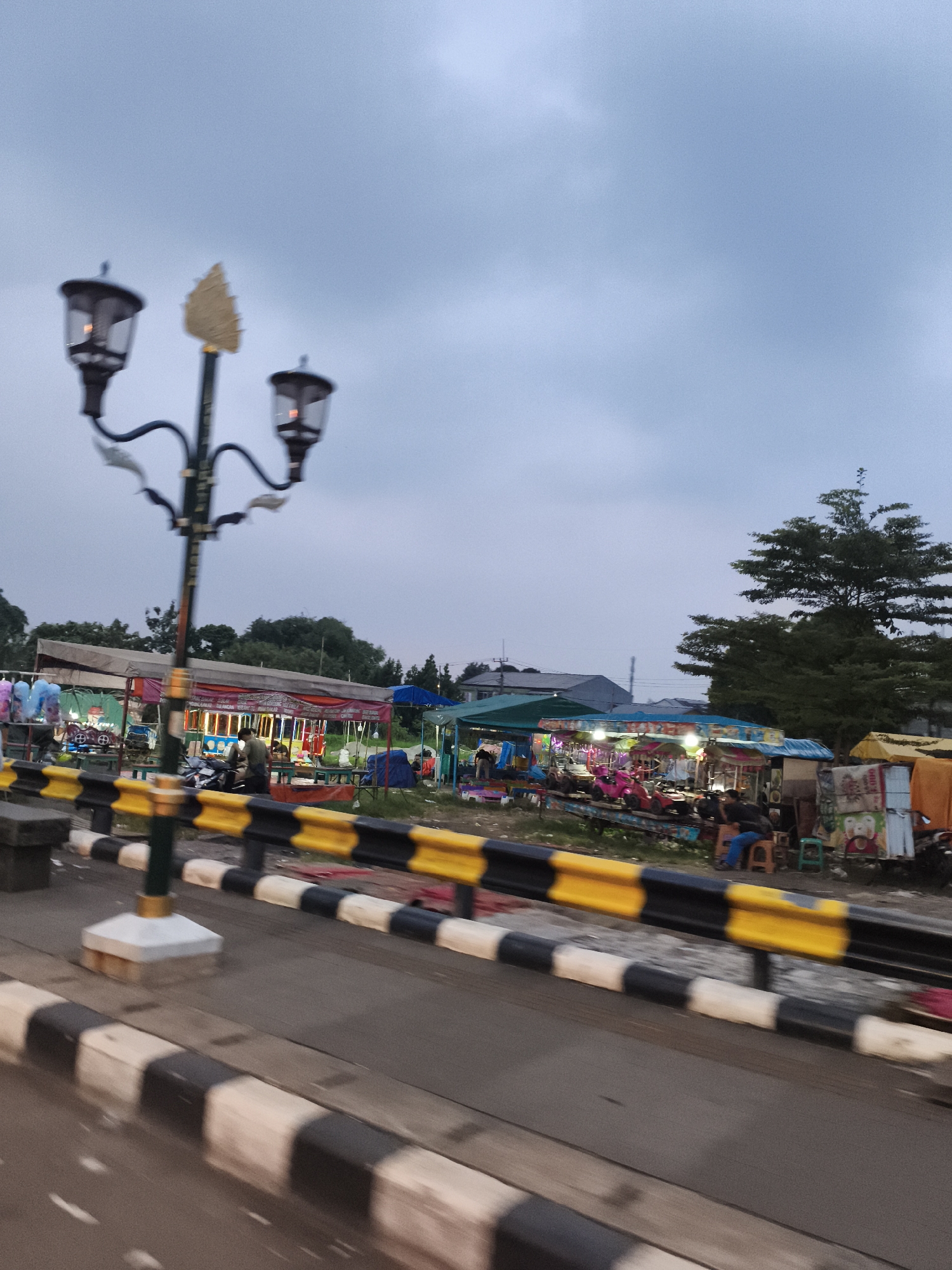 The image captures a roadside scene in Cibinong, Indonesia, likely during dusk or early evening, characterized by an overcast sky. In the foreground, a decorative street lamp with three lanterns, adorned with a golden, leaf-like finial, stands on the left. The lanterns emit a soft, warm glow.
To the right of the street lamp, a black and yellow road barrier separates the viewer's perspective (presumably from a moving vehicle, indicated by slight motion blur on the road) from a lively area. Beyond the barrier, a series of informal market stalls or small amusement rides are set up. These stalls are covered with various colored tarpaulins and tents, and some are illuminated with strings of lights, suggesting an evening market or fair atmosphere.
Several people are visible in and around the stalls. Some appear to be sitting on stools, while others are standing or tending to their merchandise. One stall features child-sized ride-on vehicles, like small cars and motorcycles, indicating it might be a children's play area or a ride. Another stall has stacked items, possibly products for sale, some with cartoon character images.
In the background, lush green trees and residential buildings with grey roofs are visible under the muted sky. The overall impression is one of a bustling local street scene transitioning from day to night, with community activity centered around the market stalls. No clearly legible text is visible in the image.