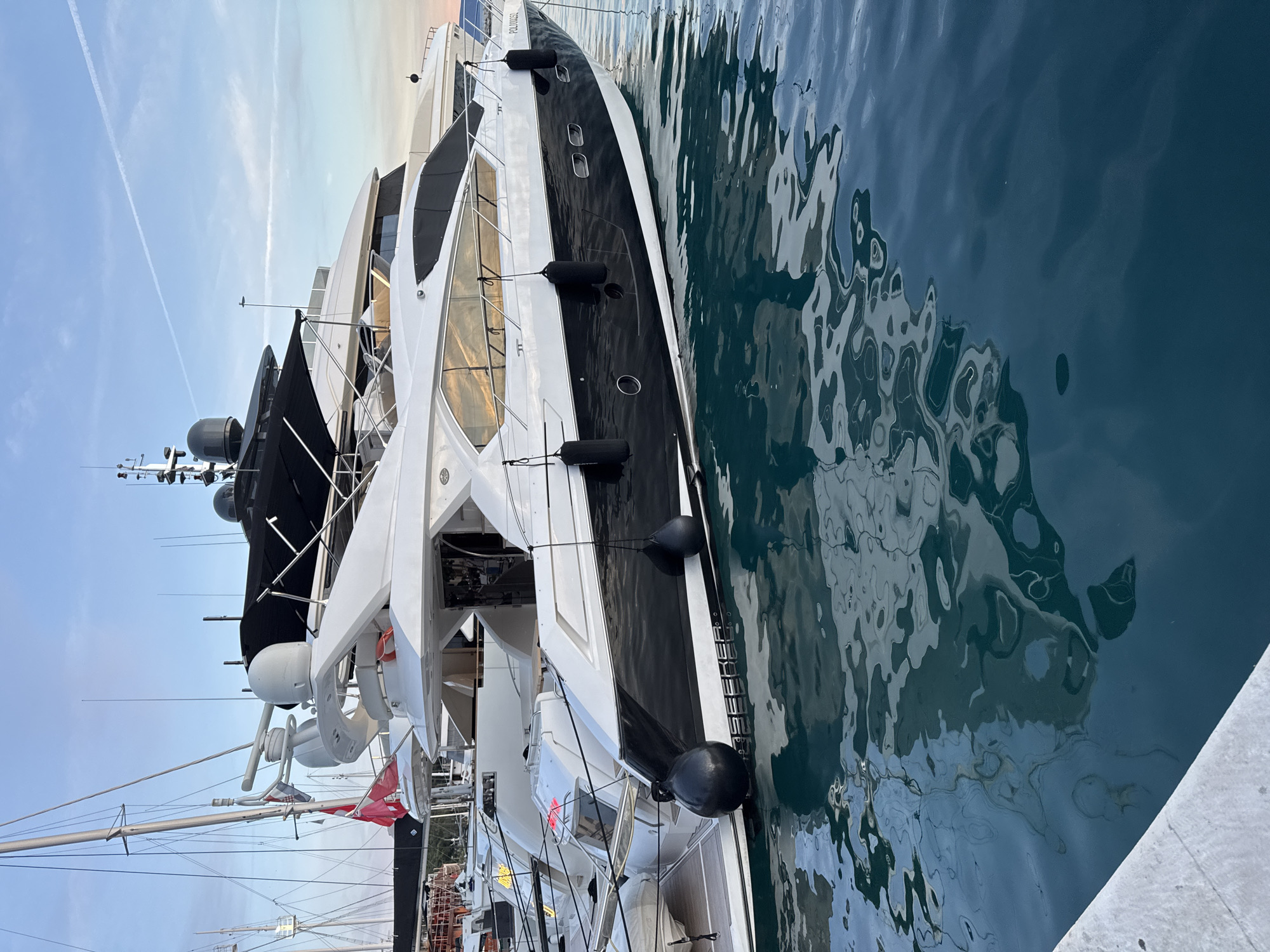 A luxurious, multi-level white and black motor yacht, identified by the name "POLO OGO" on its stern, is docked in the harbor of Makarska, Croatia. The yacht features multiple decks, large windows reflecting the sky, and black fenders attached to its hull. In the background, other sailboats and yachts are visible, along with masts and rigging. The water is a deep blue, with reflections of the yachts and the sky. The sky is a clear blue with faint white contrails. The scene appears to be captured during either sunrise or sunset, indicated by the warm hues in the sky and reflections.