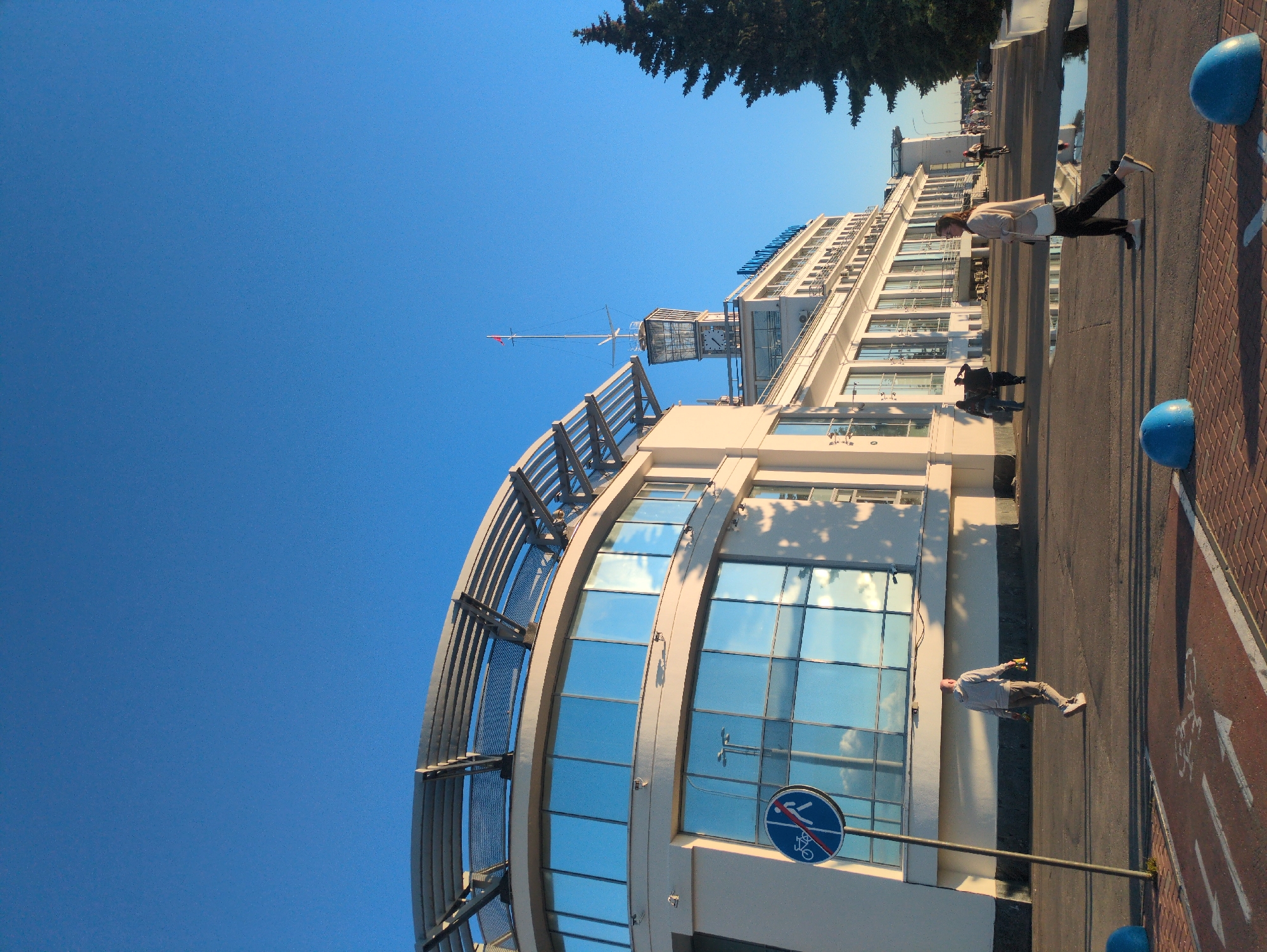 The image depicts a sunny day in Nizhny Novgorod, Russia, with a clear blue sky. The primary subject is a large, modern building with a curved, glass-fronted facade that reflects the sky and surrounding environment. The building appears to be a hotel or a similar establishment, given its scale and amenities, possibly overlooking a waterfront or a wide promenade.
In the foreground, a man is walking on a paved area, which seems to be a sidewalk or a pedestrian walkway. He is dressed casually in a light-colored shirt and trousers, carrying a yellow bag. Further in the background, another person can be seen walking away from the camera. The walkway is bordered by a red brick pavement and has white markings, including a bicycle symbol, indicating a bike path. Several rounded blue bollards are positioned along the edge of the walkway, likely for traffic control or aesthetic purposes.
A prominent feature of the building is a spire-like structure on its roof, topped with a small red flag. The building's architecture suggests a blend of modern design with some traditional elements, perhaps reminiscent of Soviet-era structures adapted for contemporary use. The sunlight casts long shadows, indicating it's either early morning or late afternoon.
A circular blue sign with a red border displays a pictogram of a bicycle with a diagonal red line through it, signifying "no cycling." This sign is mounted on a metal pole in front of the building. The overall atmosphere is one of a pleasant, public space being utilized for leisure and transit.