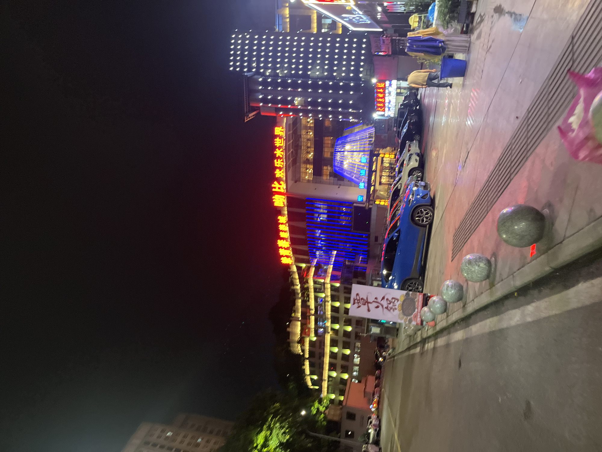This image depicts a nighttime street scene in the Jiulongpo District of China, characterized by vibrant neon lighting and commercial activity. A prominent building with a facade illuminated by numerous white lights dominates the right side of the frame, featuring an illuminated sign in red Chinese characters. To the left, another building displays a bright red neon sign with Chinese characters and is adorned with yellow and green lights on its multiple levels. In the foreground, a blue car is parked, partially obscuring a white sign with Chinese characters. Several other cars are visible on the street and in a parking area. The pavement in front of the buildings shows wet patches, suggesting recent rain. A few concrete spherical bollards are lined up along the sidewalk. A person in a yellow shirt and blue pants stands near a blue bin, while another person is partially visible in the background. The sky is dark, indicative of nighttime.