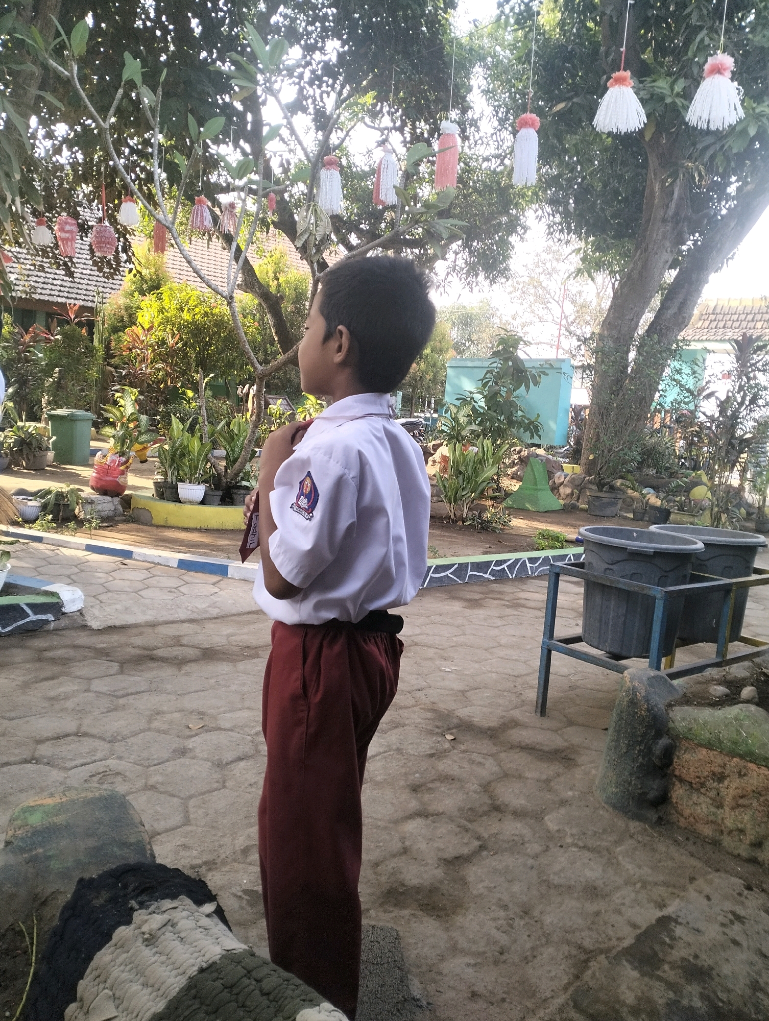The image captures a young boy standing in an outdoor area, likely a schoolyard, in Blabakan, Indonesia. He is wearing a white school uniform shirt with a school emblem on the left sleeve and dark red trousers. The boy is facing to the right, with his right hand holding the front of his shirt near his neck.
The background features lush greenery with trees and various plants. Several decorative items, resembling tassels or pompoms in white, pink, and red, are hanging from the branches of a tree. In the distance, a light blue building and a tiled roof of a building are visible. The ground is paved with hexagonal stones. To the right of the boy, there is a metal stand holding two large, dark-colored bins. Rocks and more vegetation are present in the mid-ground and background. The lighting suggests it is daytime, with sunlight filtering through the trees.