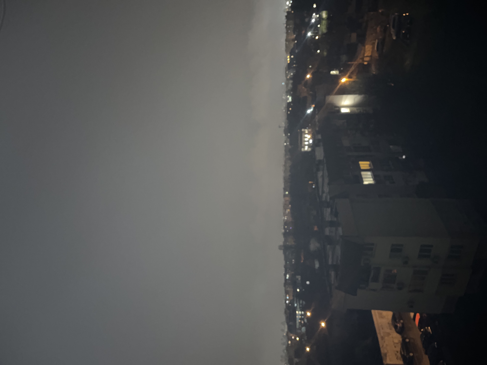 This image depicts an elevated, nocturnal view of an urban area in Sofia, Bulgaria, characterized by significant atmospheric haze or fog. The sky is uniformly grey and obscured, indicating low visibility.
In the mid-ground, a light-colored, multi-story residential building with a reddish-brown, gabled roof is partially visible on the left, presenting several illuminated windows. Adjacent to this structure, a textured, light-toned wall flanks a wet, dark asphalt surface where multiple parked cars are discernible. Several streetlights emit warm, diffused illumination, reflecting on the wet ground.
The background reveals a dense cityscape extending into the distance, marked by numerous scattered, blurred points of artificial light emanating from buildings and additional streetlights. The atmospheric haze merges with the horizon, further limiting distant detail. No individuals or specific activities are directly observable within the frame. The scene conveys a quiet, illuminated urban environment under low light and hazy weather conditions.