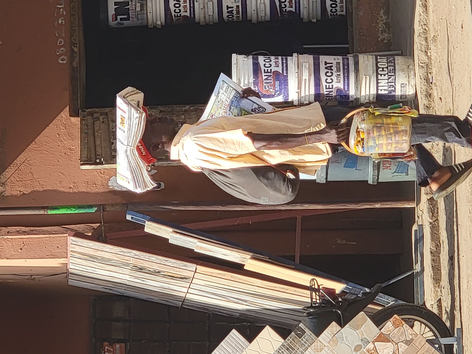 The image shows a man sitting and reading a newspaper, with stacks of newspapers next to him. He is wearing a light-colored shirt and dark pants. Next to him are several buckets of paint and a small, clear container filled with snacks or candies. In front of the man is a collection of tiles leaning against a wall and displayed on a motorcycle carrier. The background shows a building, with what appears to be a phone number painted on the side. The scene appears to be an outdoor market setting in Maiburiji, Nigeria, as suggested by the vendor's wares and general urban environment.