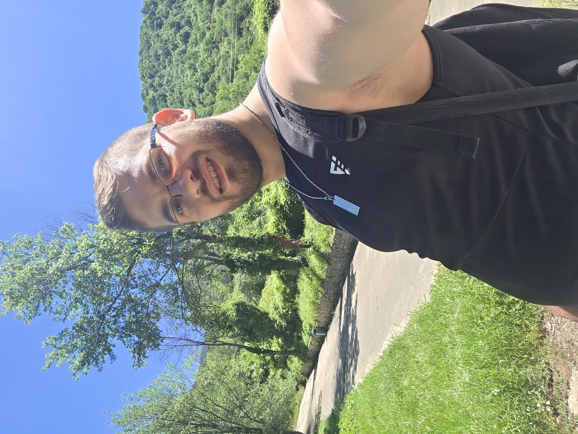 A man wearing glasses and a black shirt stands on a paved path next to a grassy area, taking a selfie. He has a beard and is wearing a silver chain with a rectangular pendant. Behind him is a green, wooded area. The photograph was taken in Bachkovo, Bulgaria.