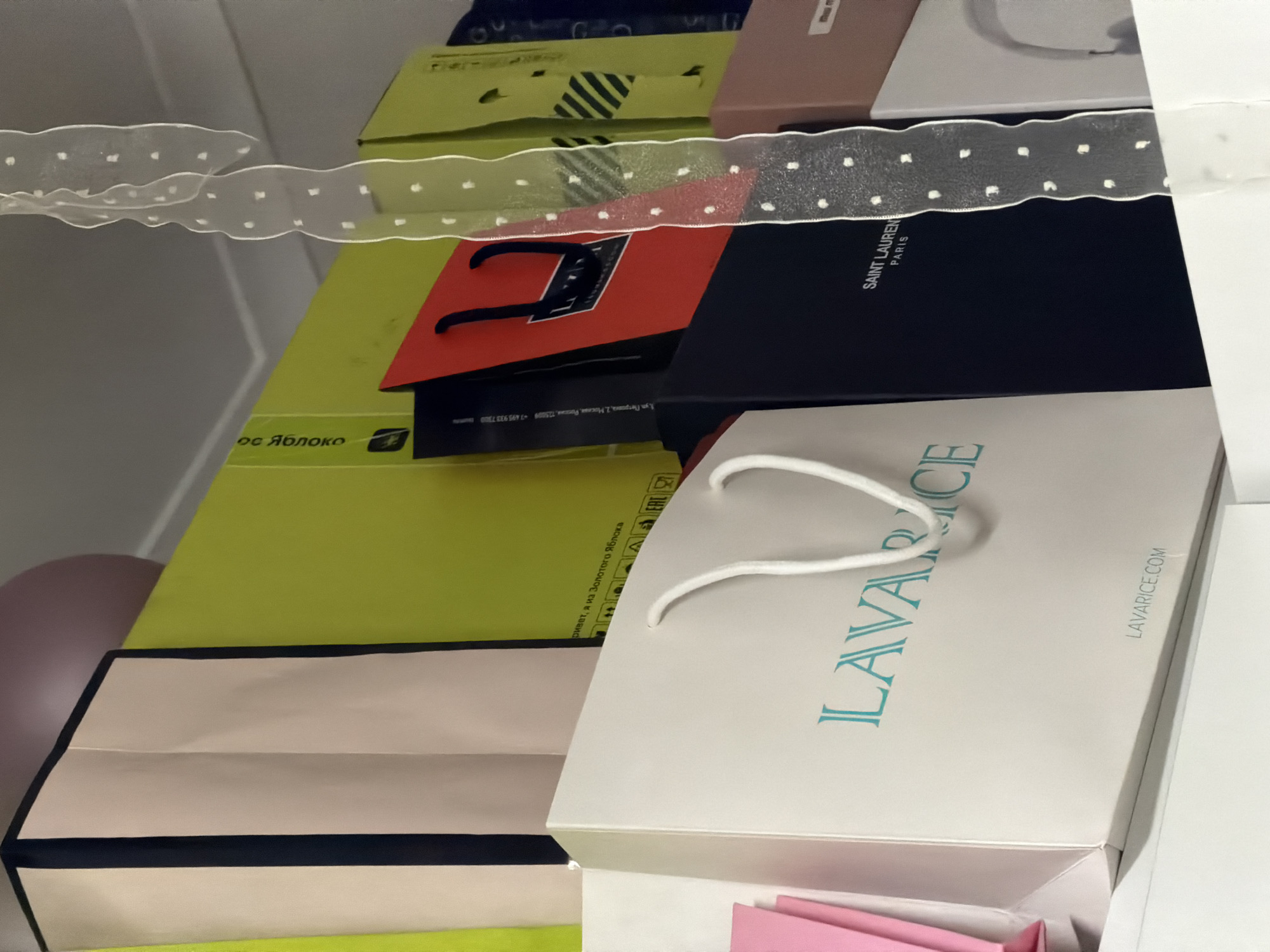 The image displays a collection of shopping bags and gift boxes stacked together on a shelf. In the foreground, a white shopping bag with blue text that reads "LAVARICE" is prominent, with the website "LAVARICE.COM" visible at the bottom. To the right of this bag is a dark navy blue box with "SAINT LAURENT PARIS" printed in white. Behind these are several other bags and boxes in various colors including lime green, pink, and red. A white ribbon with polka dots is draped vertically in the center of the image. The scene appears to be indoors, likely a closet or storage area. The text on one of the green bags, "Привет, я из Золотого Яблока", translates to "Hello, I'm from Golden Apple", which is a Russian cosmetics and fragrance retailer with stores in Moscow. This suggests the items may have been purchased from a "Золотое Яблоко" store, potentially in Moscow, Russia, given the retailer's presence there. There are no people or discernible actions depicted in the image.
