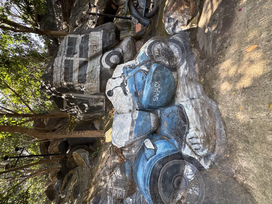 A rock sculpture depicts a blue and white motorcycle with the word "SAHARA" written on one of the blue parts. Above it, another rock sculpture has a tire and a black and white painting of what appears to be a car with the number "550" visible. The sculpture is located in an outdoor setting with trees and foliage surrounding it. The ground around the sculpture is paved.