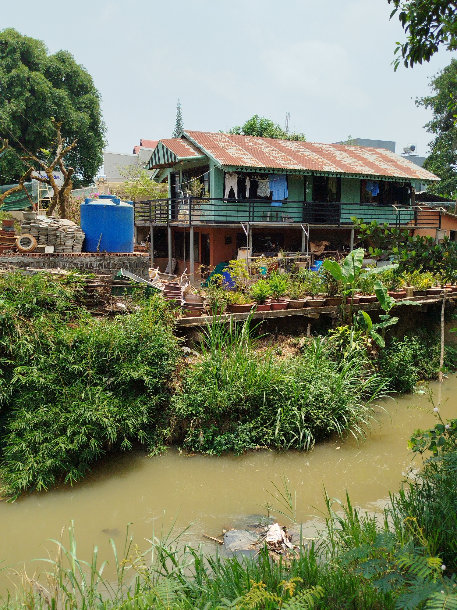The image depicts a house situated on a raised structure next to a murky canal in Tân Hiệp, Vietnam. The house is two stories with a rusted metal roof, a green exterior, and a balcony on the upper level where clothes are hanging. A blue water tank and stacked tiles are visible to the left of the house. Below the house, a variety of potted plants are arranged on a platform. The canal water is brown and contains trash. The banks of the canal are overgrown with vegetation. The sky is overcast.