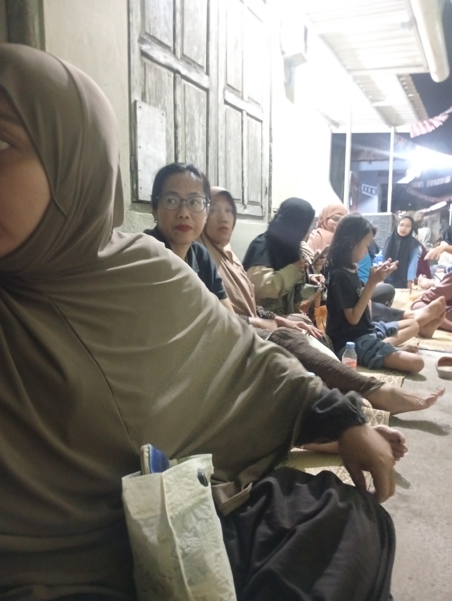The image captures a group of people, primarily women and a young girl, seated on the floor outdoors in what appears to be a residential area in Sewon, Indonesia. The foreground is dominated by the arm and shoulder of a person wearing a light brown hijab. To the left, a woman with glasses is visible, facing forward. Behind her, another woman in a beige hijab is seated. In the center and to the right, a young girl with dark hair is holding a smartphone. Several other individuals are seated around them, some partially obscured. Objects in the scene include a plastic water bottle and a light-colored patterned bag. The environment suggests a casual gathering, possibly in the evening, with a building and outdoor lighting visible in the background. The ground is paved, and there are mats on which the people are sitting.