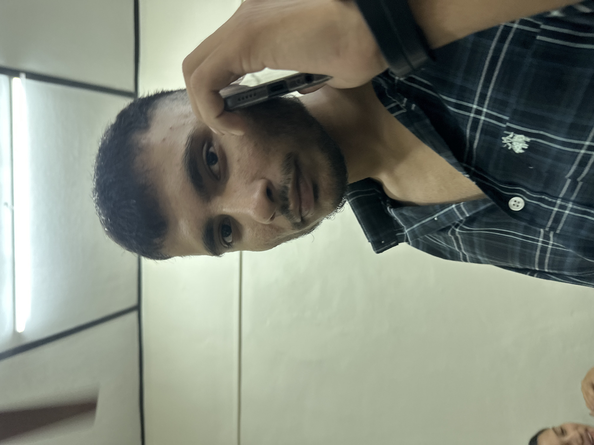 A young man is shown from the chest up, holding a smartphone to his ear as if on a call. He has short, dark hair, a mustache, and stubble. He is wearing a dark, plaid collared shirt with a white logo on the left chest. His right arm is bent, with his hand holding the phone. The background is a plain, light-colored wall with a fluorescent light fixture visible on the ceiling. The lighting is somewhat dim, creating shadows on the man's face. The image appears to be taken indoors.
