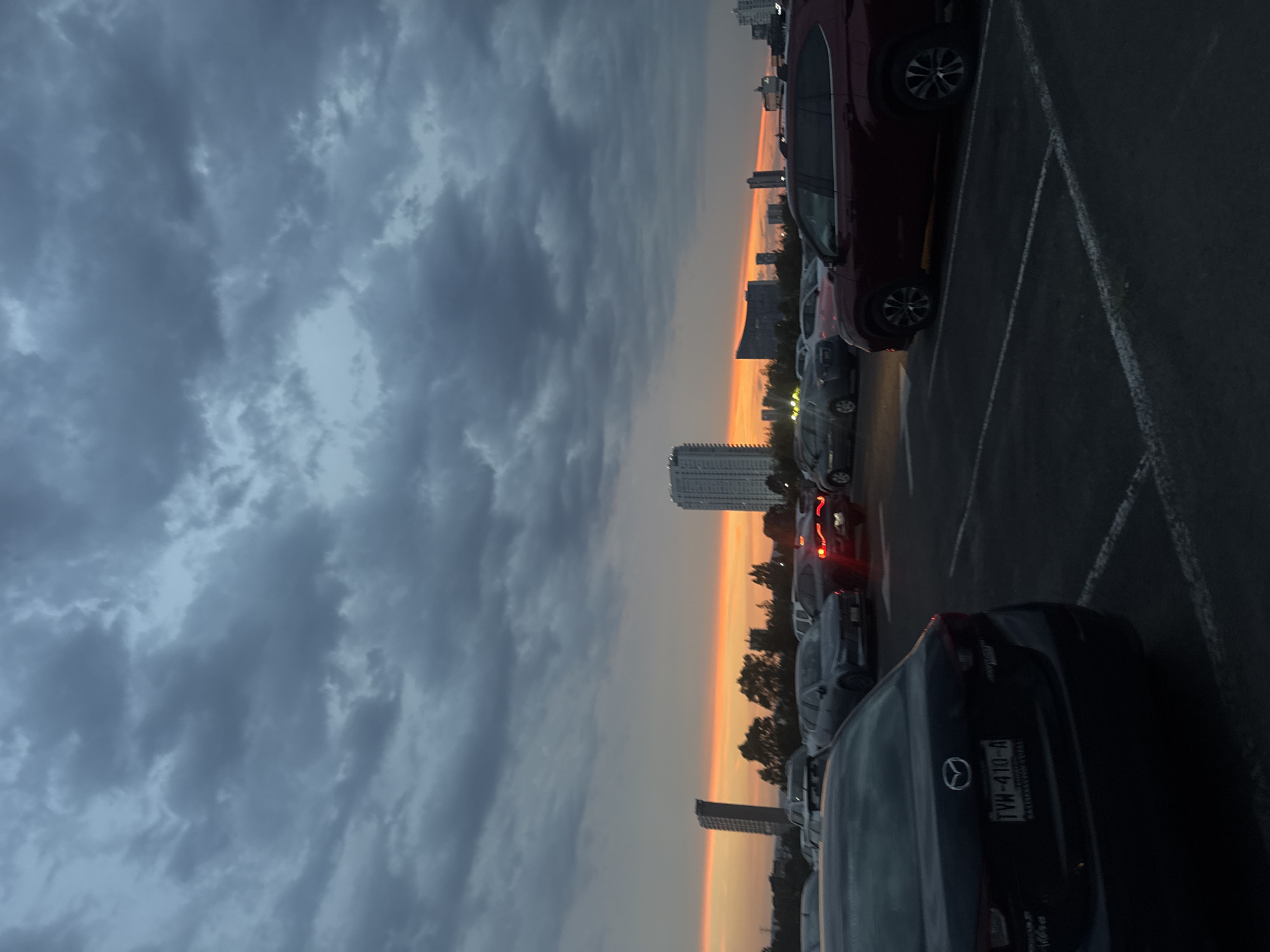 The image captures a twilight scene at a parking lot in Puebla, Mexico, with a dramatic sky. The upper and left portions of the frame are dominated by heavy, dark grey clouds, indicating an overcast sky. Along the horizon, a striking band of orange and yellow light from the setting sun cuts across the view, providing a vibrant contrast to the somber clouds. Silhouetted against this colorful horizon are several tall, modern buildings and a scattering of trees, suggesting an urban or suburban setting.
In the foreground and extending to the right, a dark asphalt parking lot is visible, marked with white lines delineating parking spaces. Numerous cars are parked, mostly dark-colored. A prominent dark red car, likely an SUV or crossover, is seen parked on the right side. In the immediate foreground, the rear of a dark blue or black Mazda is clearly visible, featuring the Mazda logo and a license plate that reads "TVW-410-A," with "Puebla" inscribed beneath the numbers, confirming the location. Further into the parking lot, faint red glows from some cars' brake lights are discernible. The overall atmosphere is one of transition, as the last light of day fades into night.
