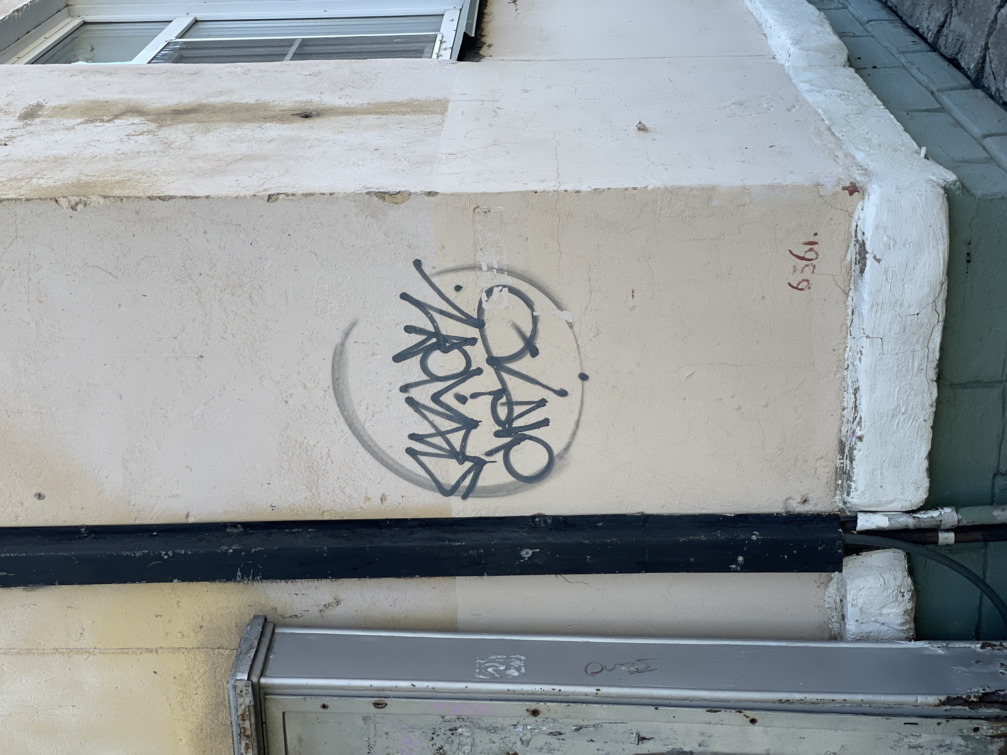 The image displays the exterior corner of a weathered building in Zheleznogorsk, Russia. The primary wall is painted a light creamy-beige, exhibiting cracks, peeling paint, and general discoloration. Centered on this wall, a grey spray-painted graffiti tag, consisting of stylized, illegible lettering, is enclosed within an incomplete grey circular outline. Below the graffiti, closer to the ground, the red hand-painted numbers "6361." followed by a dot are visible.
To the left, a portion of a grey metal utility box or panel, showing rust and wear, is partially visible. A vertical black conduit or downpipe runs along the junction of the utility area and the beige wall, displaying chipped paint and surface wear. This pipe connects to smaller, darker conduits at the base of the wall.
The adjacent wall, to the right of the corner, is painted a lighter off-white and shows significant water staining and wear, particularly vertical streaks below a partially visible white-framed window.
The base of the building features a prominent white-painted concrete or plaster band, which is cracked and textured. Beneath this band, the foundation consists of dark teal or green painted bricks or blocks. The ground adjacent to the foundation appears to be paved with cobblestones or irregular stone slabs. No individuals are present in the scene.