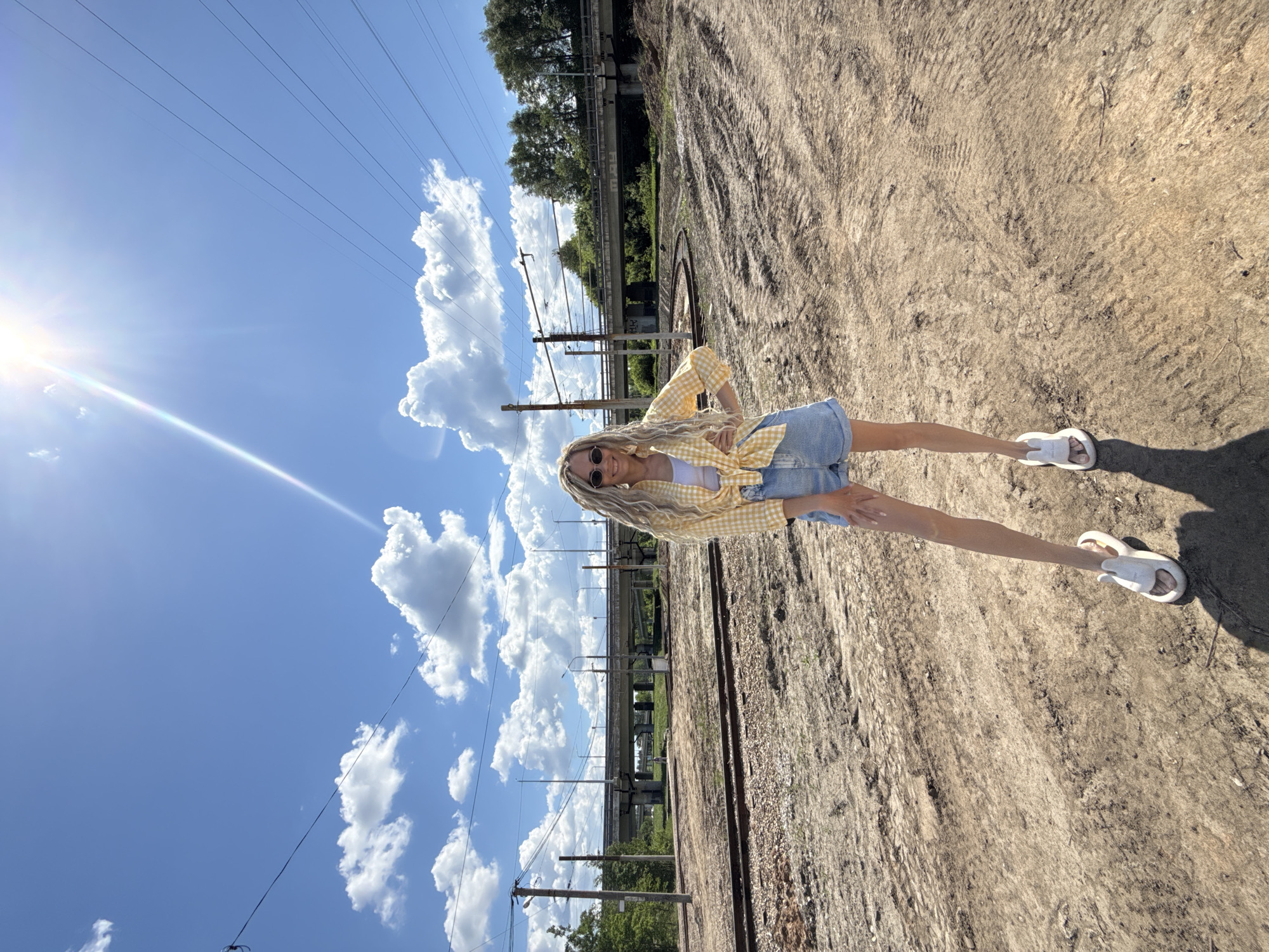 A young woman with blonde dreadlocks is standing on a set of train tracks in an open, sunny area. She is wearing a white tank top, a yellow-and-white checkered button-down shirt, a denim skirt, and white sandals. She is posing with her hands on her hips, looking towards the camera, and wearing dark sunglasses. The environment includes a bright blue sky with puffy white clouds, visible power lines, trees, and a bridge or overpass in the background. The ground is covered in dry dirt and gravel. The sun is shining brightly, causing a lens flare in the upper left corner of the image. The image location is Заволжье, Russia.