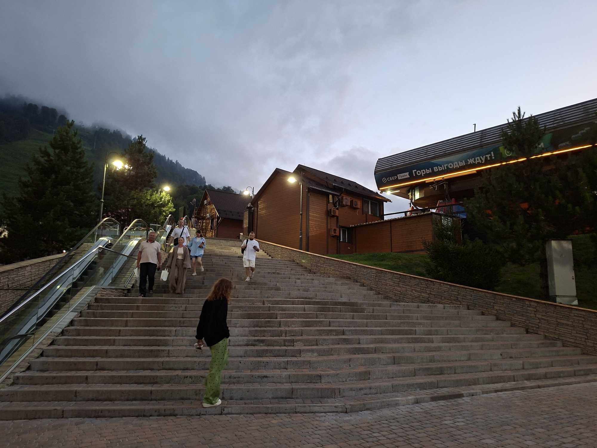 In the city of Estosadok, Russia, a wide, stone staircase ascends towards a ski lift station. The sky is overcast with a muted twilight glow. Several people are walking up the stairs. A woman in a black jacket and green polka-dot pants walks away from the camera. A man in a white polo shirt and shorts walks down with a shopping bag. Further up the stairs, a woman in a white top and beige skirt walks with another woman in a light blue shirt and dark skirt. A man in a light-colored shirt walks past them. To the left of the stairs, an escalator is visible, also with people on it. The ski lift station, with its signage and illuminated edges, is positioned at the top of the rise. Wooden buildings and pine trees are situated on the slopes behind the staircase. The ground is paved with brickwork.