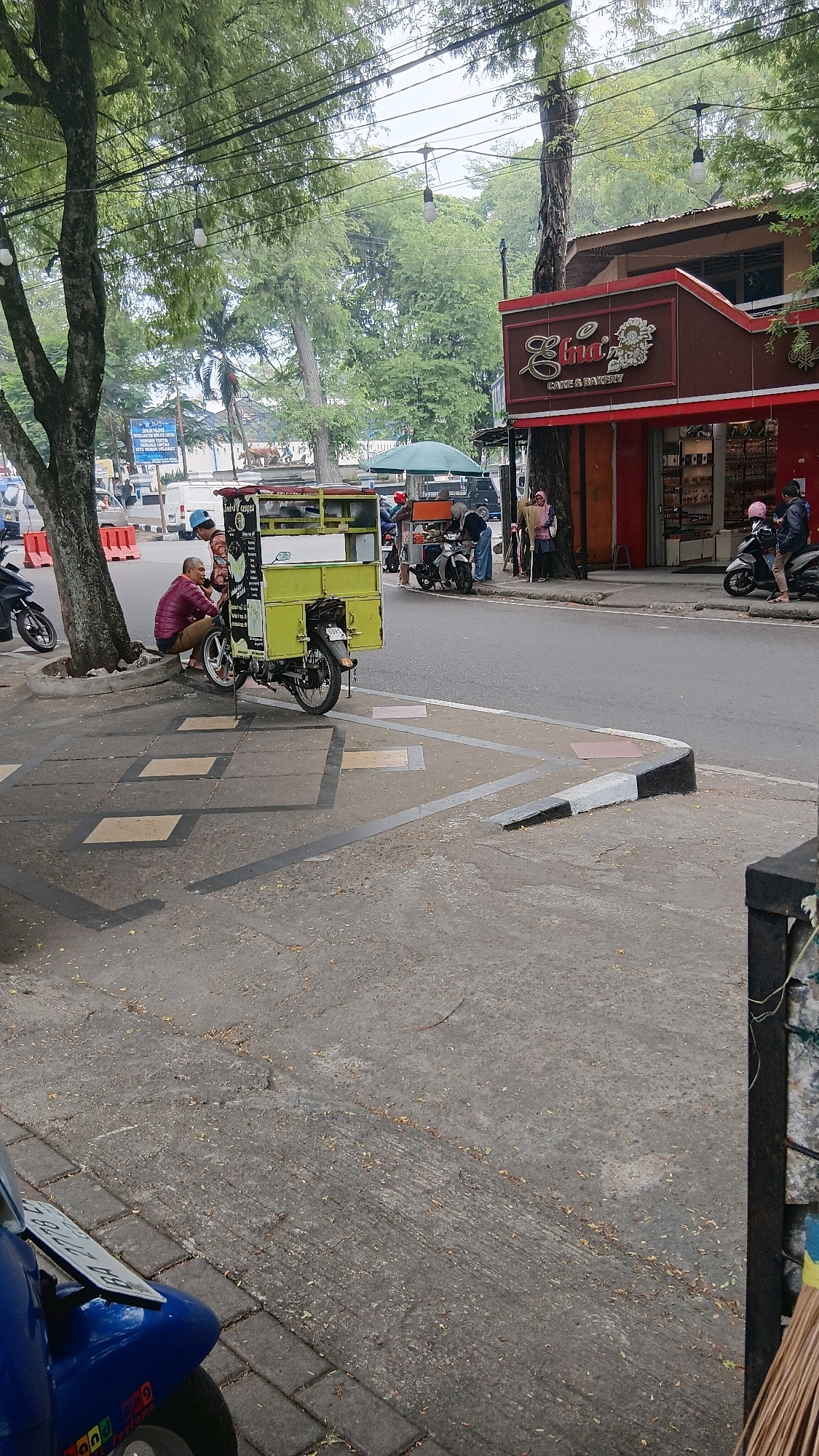 A street scene in Baringin, Indonesia, shows a sidewalk lined with trees and a street. On the sidewalk, a man in a maroon jacket and brown pants is seated next to a bright yellow cart attached to a motorcycle. Two other individuals are near the cart, one possibly engaged in conversation with the seated man. The cart appears to be a food vendor. Further down the sidewalk, several people are gathered around motorcycles, and a pink-roofed building with a sign reading "Ethi Cake & Bakery" is visible. Traffic and other parked vehicles can be seen in the background, suggesting a moderately busy urban environment. The sidewalk is paved with concrete and decorative tiles, and a marked pedestrian crossing is present near the curb.