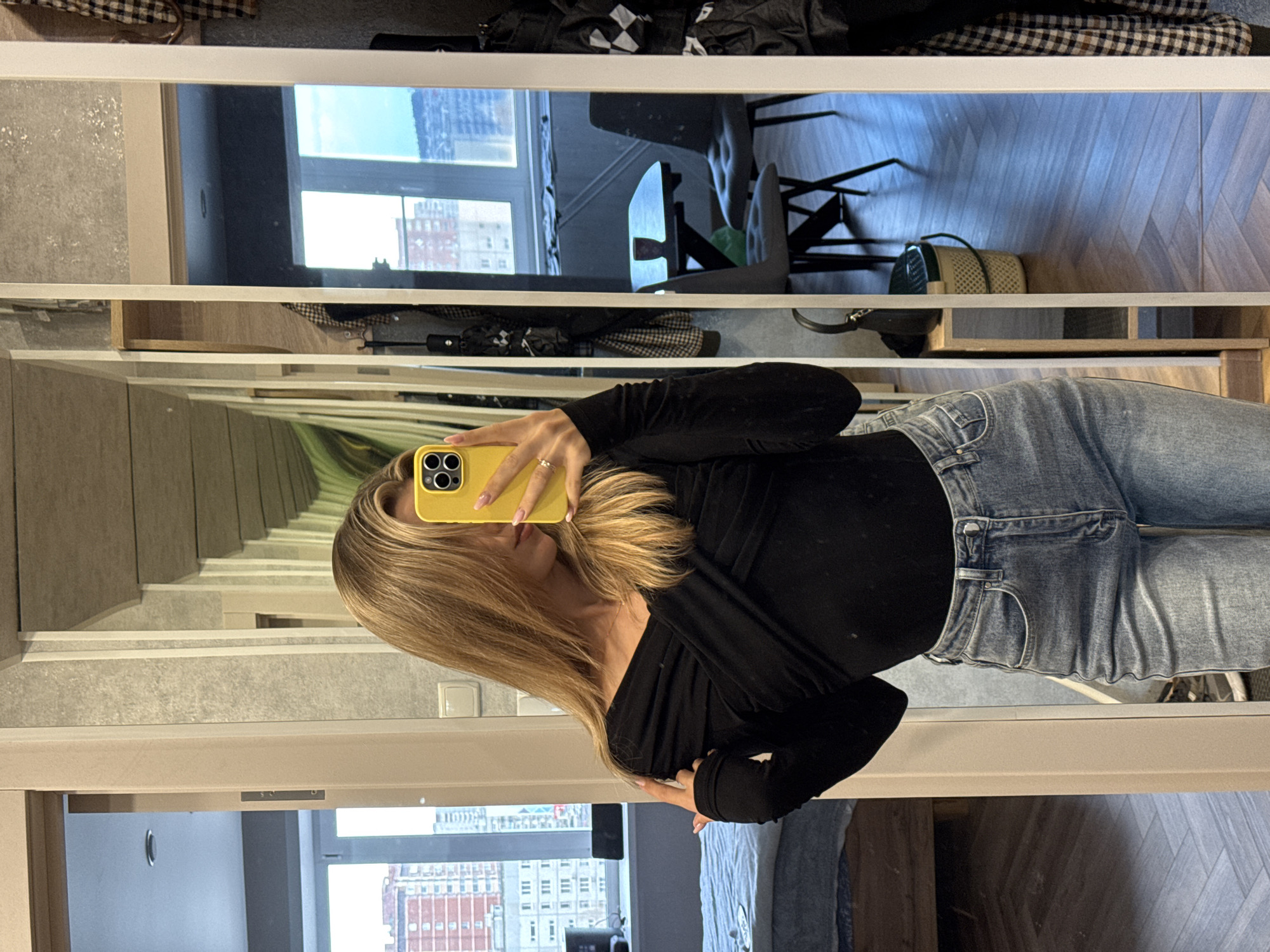 A woman is taking a selfie in a mirror. She is wearing a black top and blue jeans. Her hair is blonde and long. She is holding a yellow iPhone. The mirror is in a room with a window that overlooks a city. The city has tall buildings and a blue sky. The room also has a chair, a table, and other objects that are reflected in the mirror.