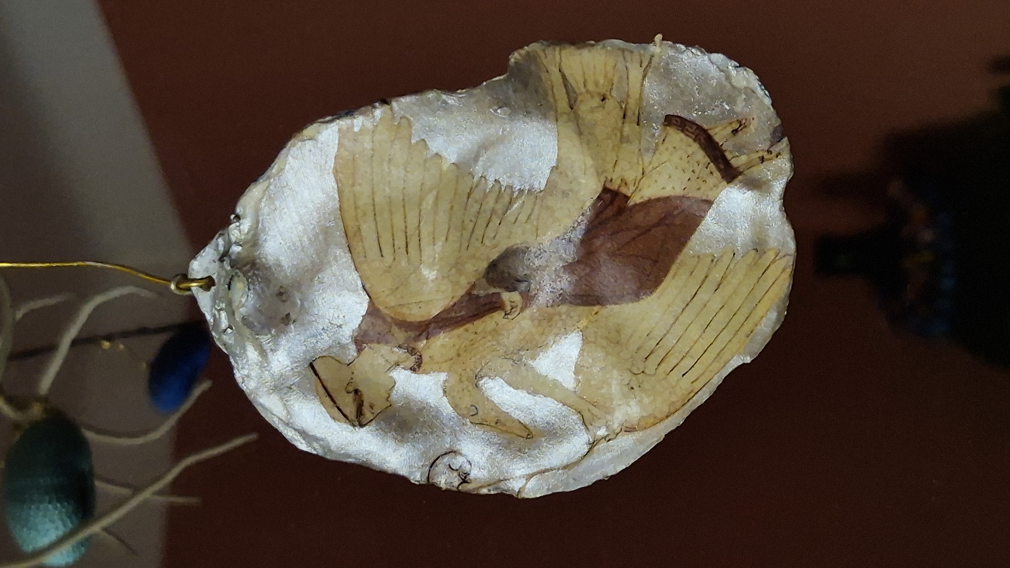 This is a close-up, vertically oriented photograph of a unique pendant or ornament, likely made from a natural material such as a shell or a piece of wood, that has been artfully decorated. The ornament is oval-shaped with an irregular edge and appears to be coated in a translucent resin. Embedded within or painted onto this material is a stylized depiction of a winged figure. The figure has a distinct profile with a headdress, and its body is rendered in muted tones of yellow, beige, and brown, suggesting ancient or classical artwork. The wings are prominent, depicted with thin, parallel lines that mimic feathers. The background is a rich, warm brown, possibly a wall or a fabric. Above the ornament, out of focus, are other decorative elements: a textured teal bead and a small blue bead, suspended by thin wires. The overall impression is of a handcrafted, decorative item. There is no discernible information or context within the image that places it in Taranto, Italy.