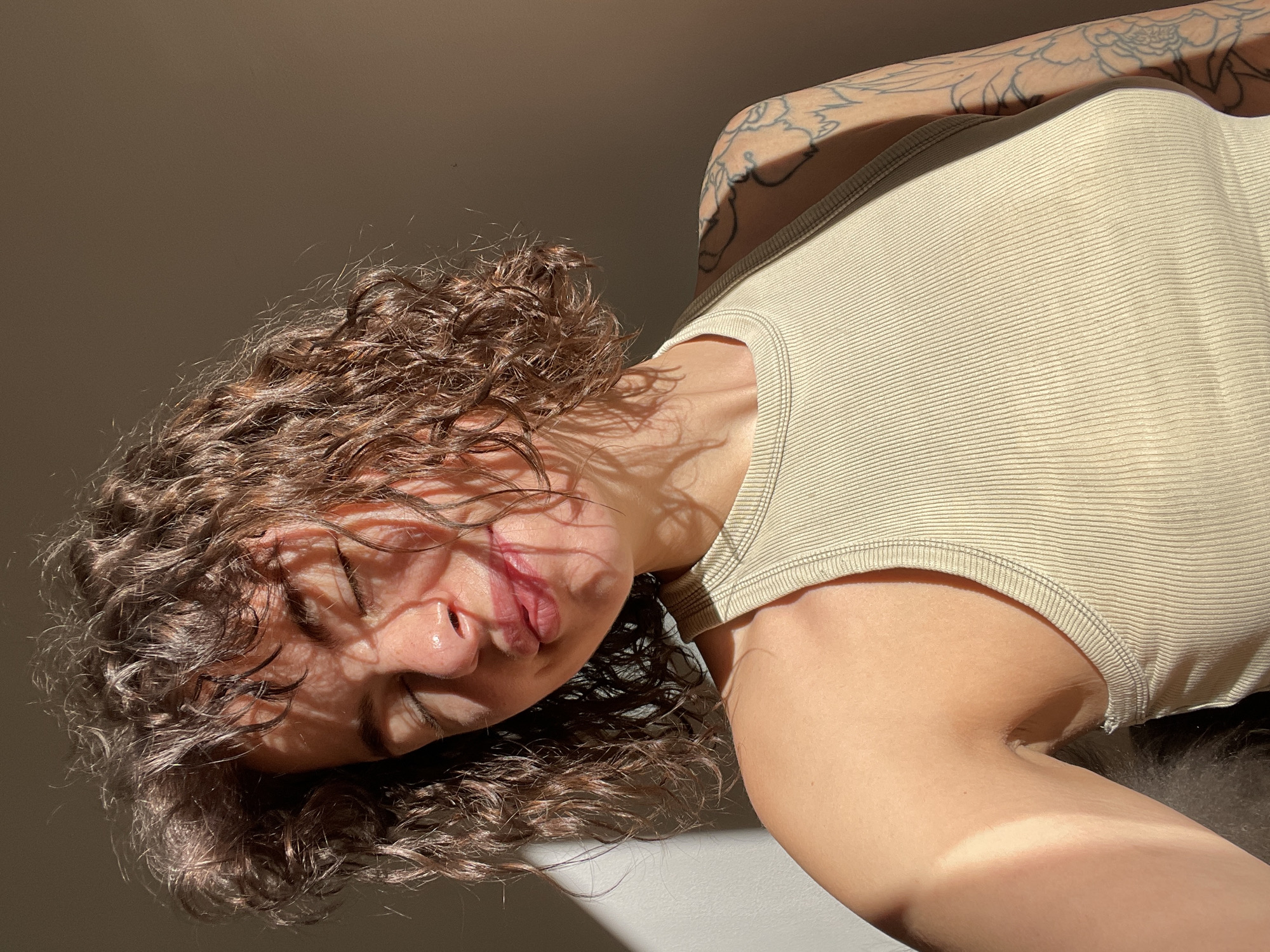 An intimate, close-up portrait features a person with abundant, dark brown curly hair, depicted in Boreti, Montenegro. The shot is angled, presenting the subject with their head tilted and eyes closed or looking downwards, conveying a sense of repose or contemplation. They are wearing a light beige, ribbed tank top. Intricate floral or leafy tattoos are visible on their left arm and shoulder.
The image is characterized by strong, directional natural light, likely from early morning or late afternoon sunlight (golden hour). This light casts striking, elongated shadows across the person's face, neck, and body, creating abstract patterns that enhance the visual texture. The sunlight beautifully highlights individual curls of the hair, giving it a rich, lustrous appearance. The skin, illuminated by the warm glow, contrasts with the distinct shadow areas. A visible tan line can be seen on their right arm and shoulder. The background is a plain, light-colored surface, maintaining focus on the subject and the dramatic interplay of light and shadow. The overall mood is serene and peaceful. No text is visible in the image.