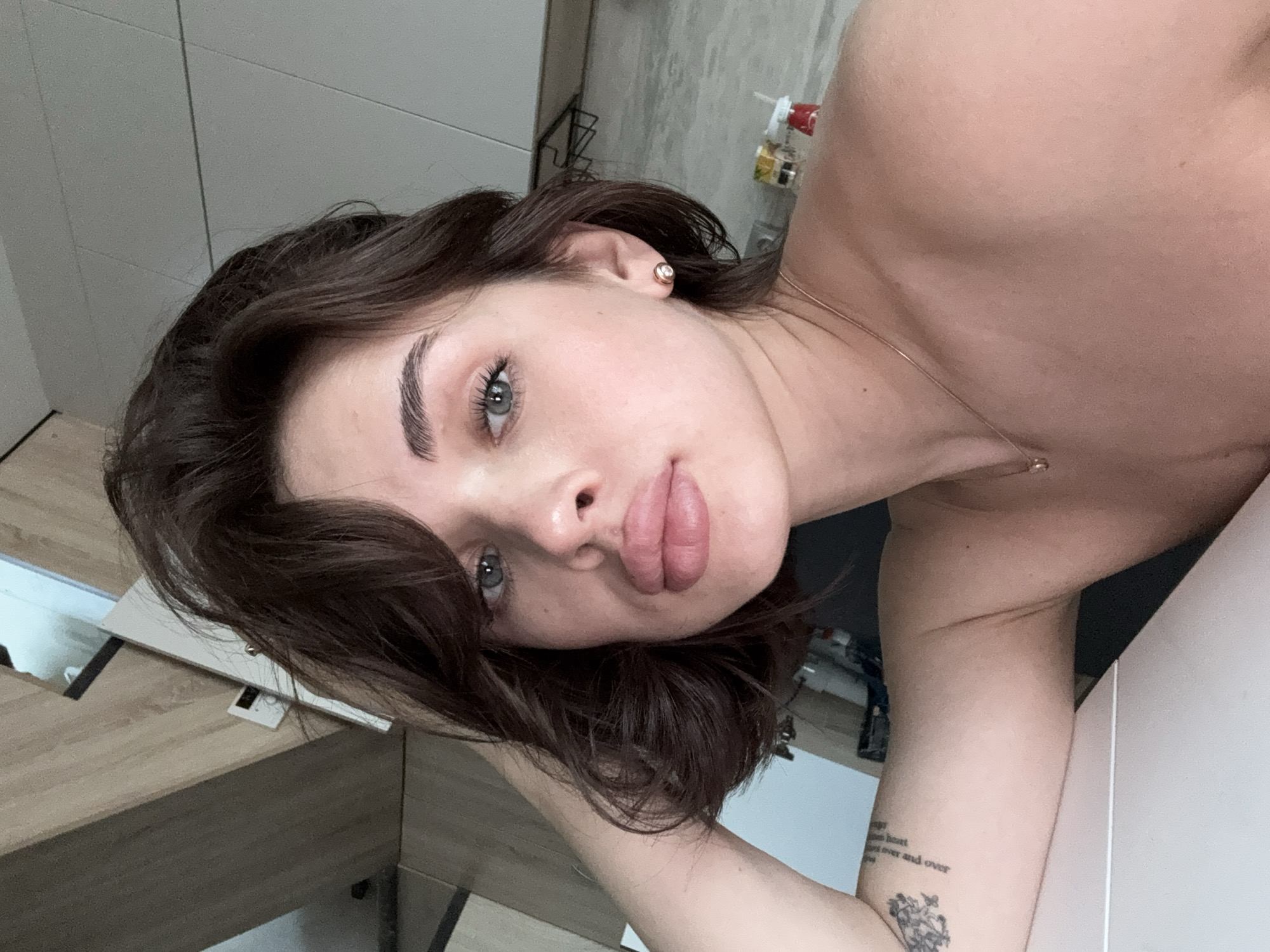 A young woman with brown hair and light eyes is positioned close to the camera, her head tilted. She is wearing a small earring in her left ear and a delicate necklace. A tattoo is visible on her left forearm. The background includes a countertop and a cabinet in what appears to be a bathroom or kitchen. The location is Лобаново, Russia.