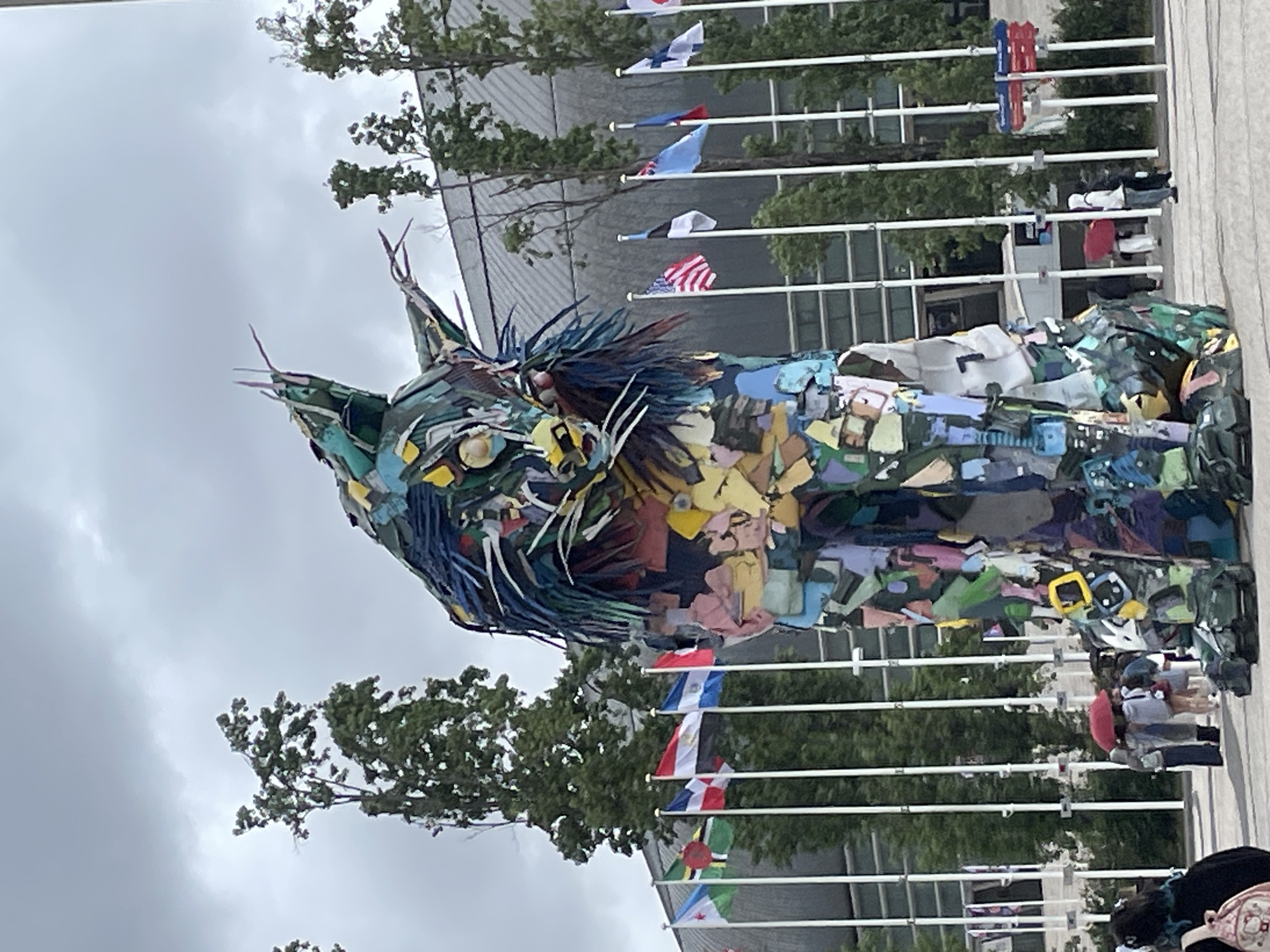 The image depicts a large, colorful sculpture of a lynx, seemingly made from recycled materials. The sculpture stands in front of a building with numerous flagpoles displaying various national flags, including the US flag. People are walking around the area. The sky is overcast. Based on the sculpture's location and design, it is likely located in Lisbon, Portugal, near the United Nations headquarters building.