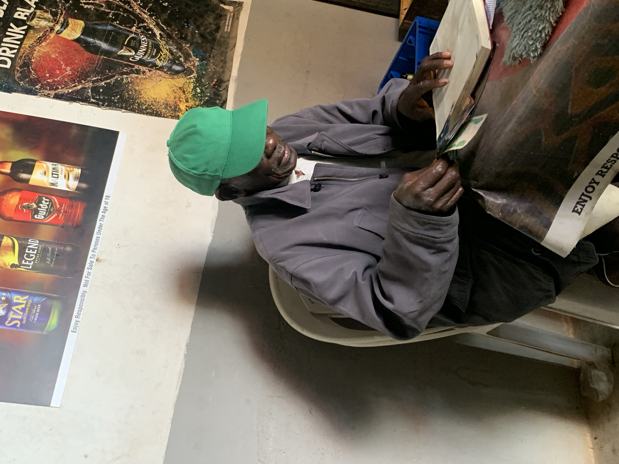 The image captures an indoor scene, likely in a shop or establishment, with promotional posters for alcoholic beverages adorning the wall in the background. In the foreground, an elderly Black man wearing a grey jacket and a green baseball cap sits at a table. His hands are engaged with what appears to be a small stack of bills and an open book or ledger. The table is covered with a dark, patterned material, and a blue plastic crate is visible to the right. The environment appears somewhat rustic, with visible wear on the walls and floor. The man's posture and the presence of money and a ledger suggest he might be involved in a transaction or record-keeping. The promotional posters depict brands such as "Star," "Legend," "Golder," "Maltina," and "Guinness," with text related to responsible drinking. The overall impression is that of a local business setting in Nigeria.