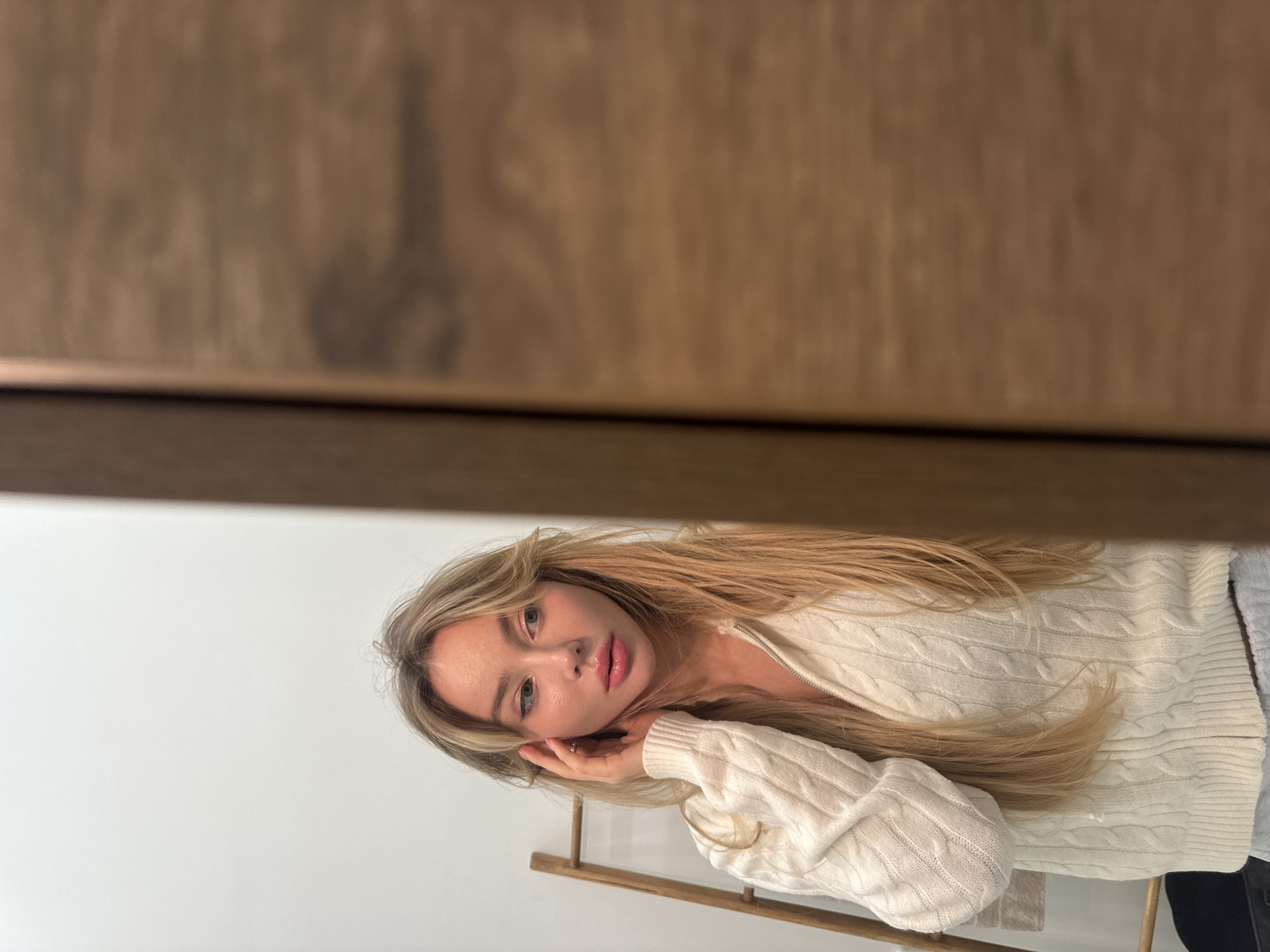 A woman with long blonde hair poses for a photograph. She is wearing a cream-colored cable knit sweater. Her face is tilted, and her hand is near her cheek. A light-colored wall is in the background, along with a wooden structure. The image has a wooden frame or surface partially obscuring the view from the top. There is no indication of Moscow, Russia.