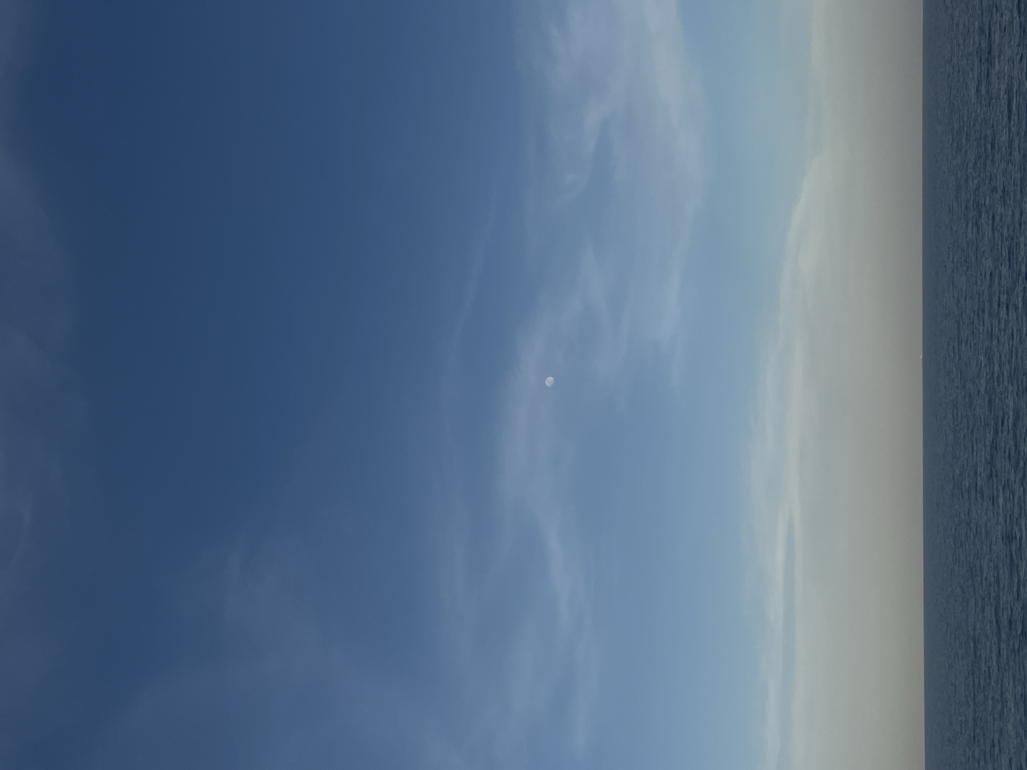 The image shows a light blue sky with wispy clouds and a very small, pale moon visible. The horizon line is on the right side of the image, with a dark blue body of water visible. The location is in the city of Unknown, Unknown. There are no people, objects, or actions occurring in the scene beyond the natural elements.