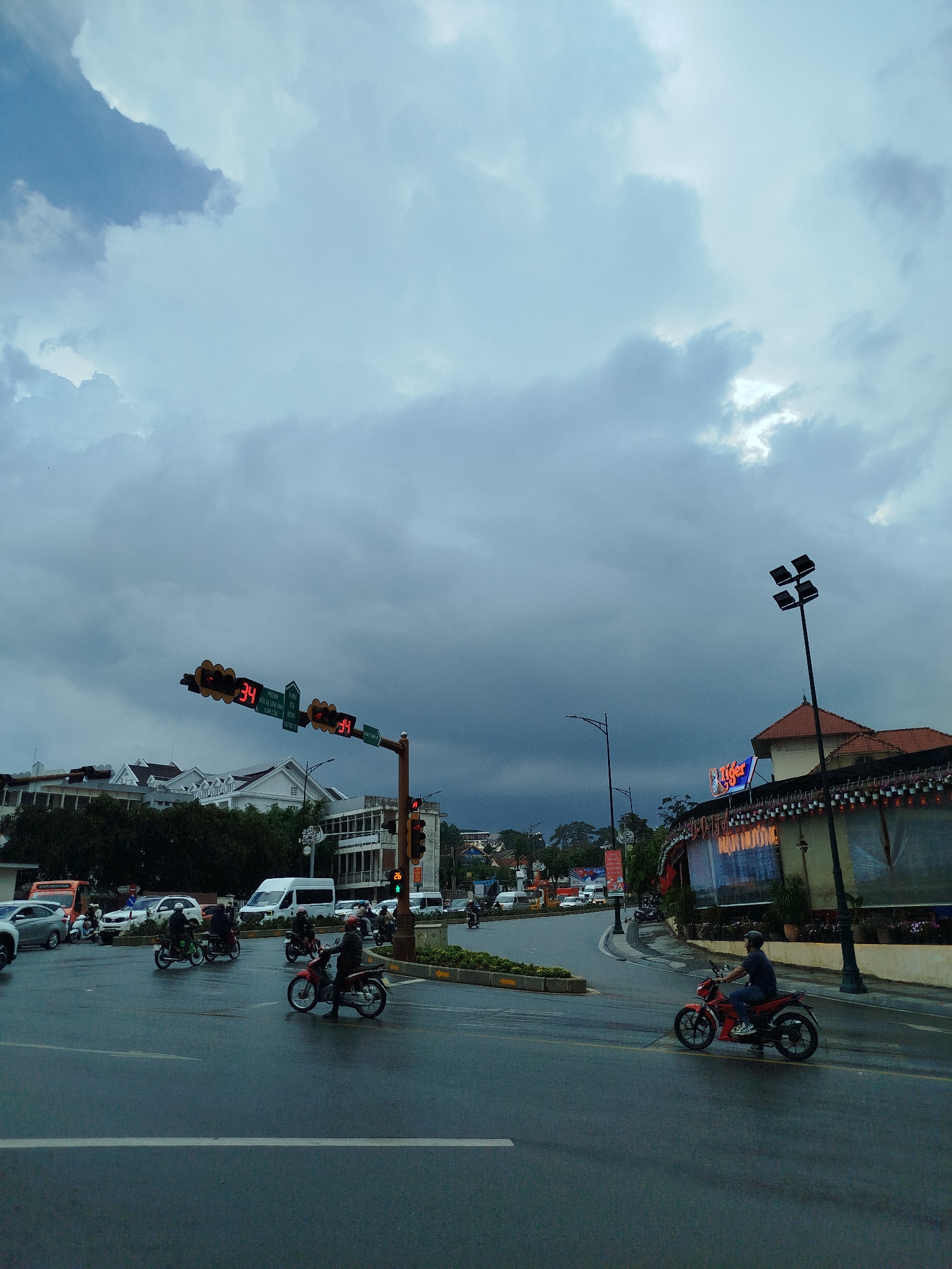 The image depicts a street scene in Da Lat, Vietnam, characterized by a wet road surface indicative of recent rain. Several motorbikes are visible in the intersection, some in motion and others stationary, waiting for traffic signals to change. The traffic lights display countdown timers alongside street names in Vietnamese. Buildings flank the intersection, including a multi-story structure with a white facade and a restaurant adorned with decorative lights and a large "Tiger" beer sign. The sky is overcast with heavy, gray clouds. A utility pole with multiple lights stands on the right side of the image. Cars and other vehicles are visible in the background, suggesting moderate traffic flow. People on the motorbikes are wearing helmets, and their clothing appears typical for everyday street attire.