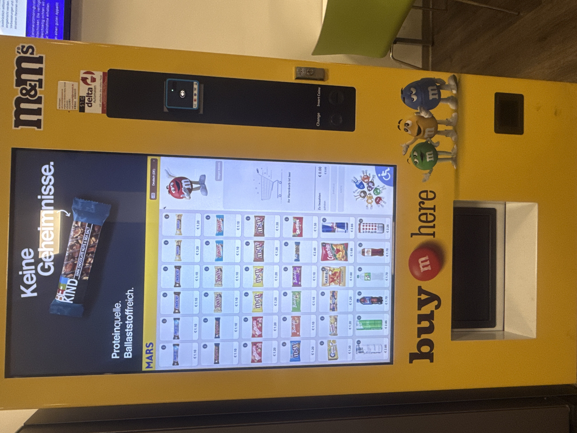 A brightly lit, yellow M&M's branded vending machine stands indoors, likely in a public or office setting, indicated by a partial view of a green chair and a wooden floor in the background. The machine is designed with M&M's characters (blue, yellow, and green) acting as mascots on its upper right side. The large touch screen interface displays various snacks and drinks for sale, with their respective prices in Euros.
Prominently featured on the left side of the screen is a "BE KIND DARK CHOCOLATE NUTS & SEA SALT" bar, alongside text in German: "Keine Geheimnisse." (No secrets.) and "Proteinquelle. Ballaststoffreich." (Protein source. Rich in fiber.) and "MARS". The main display shows a selection of items including Snickers, Twix, Maltesers, M&M's, Balisto, Skittles, Haribo, Crunchips, and various beverages like Red Bull, Coca-Cola, and water brands.
The screen currently reads "Ihr Warenkorb ist leer" (Your shopping cart is empty) and "Zu bezahlen € 0.00" (To pay € 0.00), suggesting no items have been selected yet. Options for payment methods like "BARGELD" (Cash) and "KARTE" (Card) are visible. Above the screen, there are slots labeled "Change" and "Insert Coins", as well as a small screen possibly for card payments. A "delta Automaten" logo is visible, indicating the vending machine operator, with the slogan "Wir wünschen einen guten Appetit!" (We wish you a good appetite!). Below the M&M's characters, the machine has a dispensing slot and reads "buy m here" with a large red M&M's logo. Text related to feedback ("arianeratzmann@cati", "Verbesserungen", etc.) is also partially visible on the upper left screen. The language setting is "Deutsch (DE)".