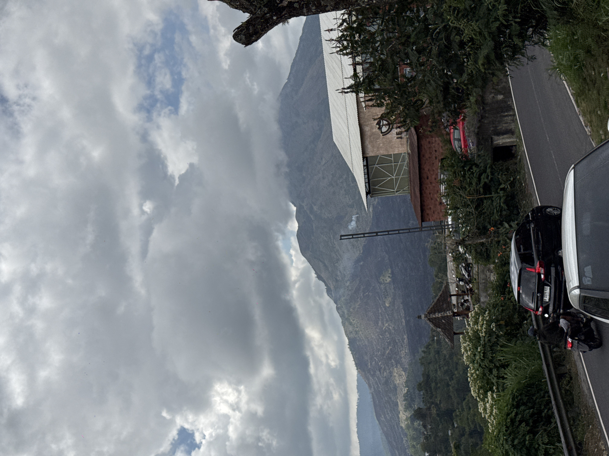 In Kintamani, Indonesia, a winding asphalt road curves through a lush green landscape leading towards a majestic, mist-shrouded volcano. The sky is overcast with dramatic, heavy clouds. On the right side of the road, a multi-story building with a corrugated metal roof stands prominently, surrounded by dense foliage. Further down the road, a traditional Balinese gazebo with a thatched roof offers shelter to several parked motorcycles. A black SUV is stopped on the road, its brake lights illuminated, and a motorcyclist is behind it. In the foreground, the hood of a white car is visible. The slopes of the volcano exhibit a dark, barren appearance, hinting at its volcanic nature.