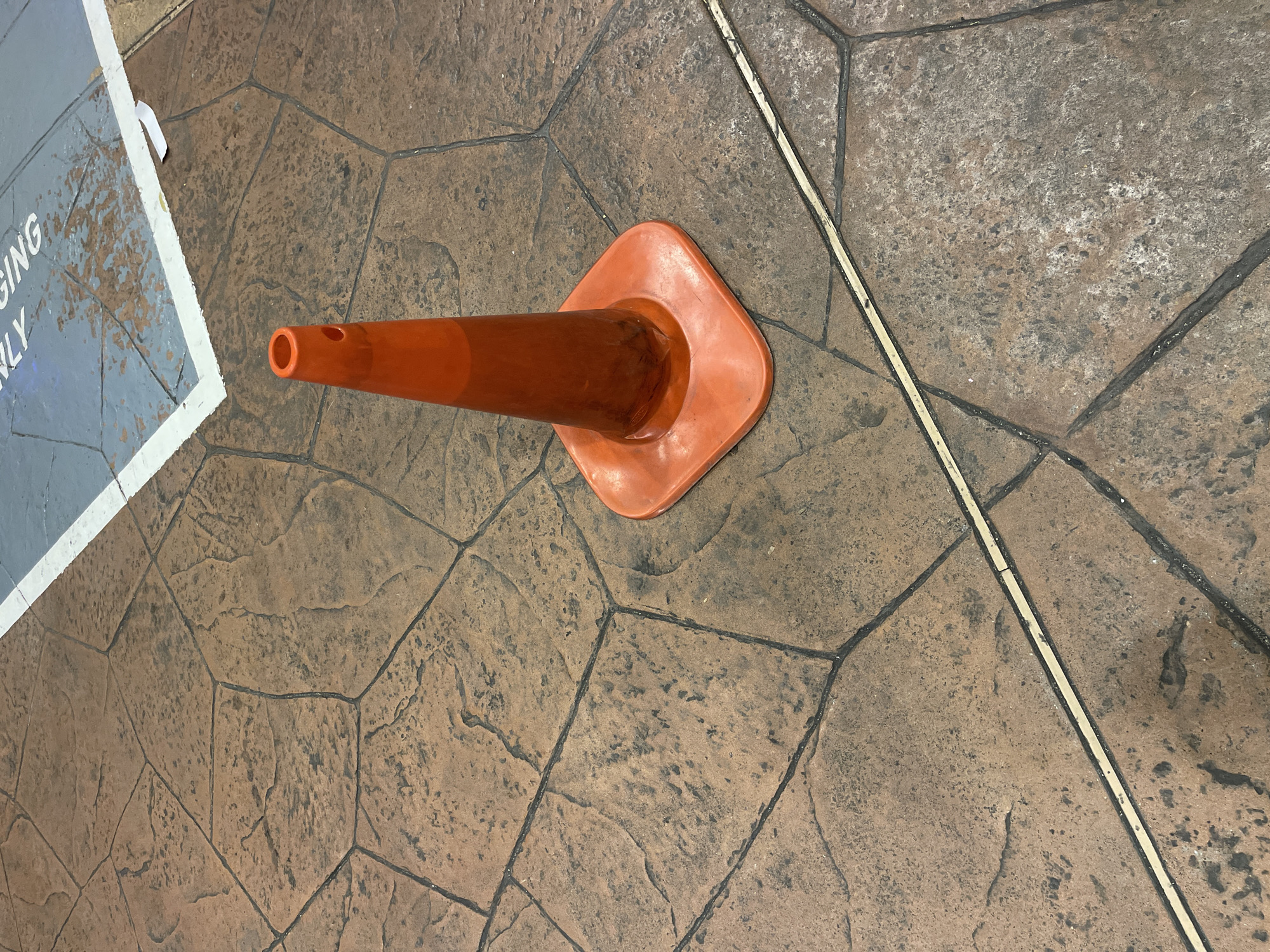 A bright orange traffic cone stands on a patterned stone floor, a section of a blue painted surface with white markings is visible in the upper right corner. The stone floor has a reddish-brown hue and is divided by dark grout lines forming an irregular pattern. The blue surface appears to have some scuff marks. The image was taken from a slightly elevated angle, looking down. The location is not identifiable as Kuala Lumpur, Malaysia based on the visual information provided.