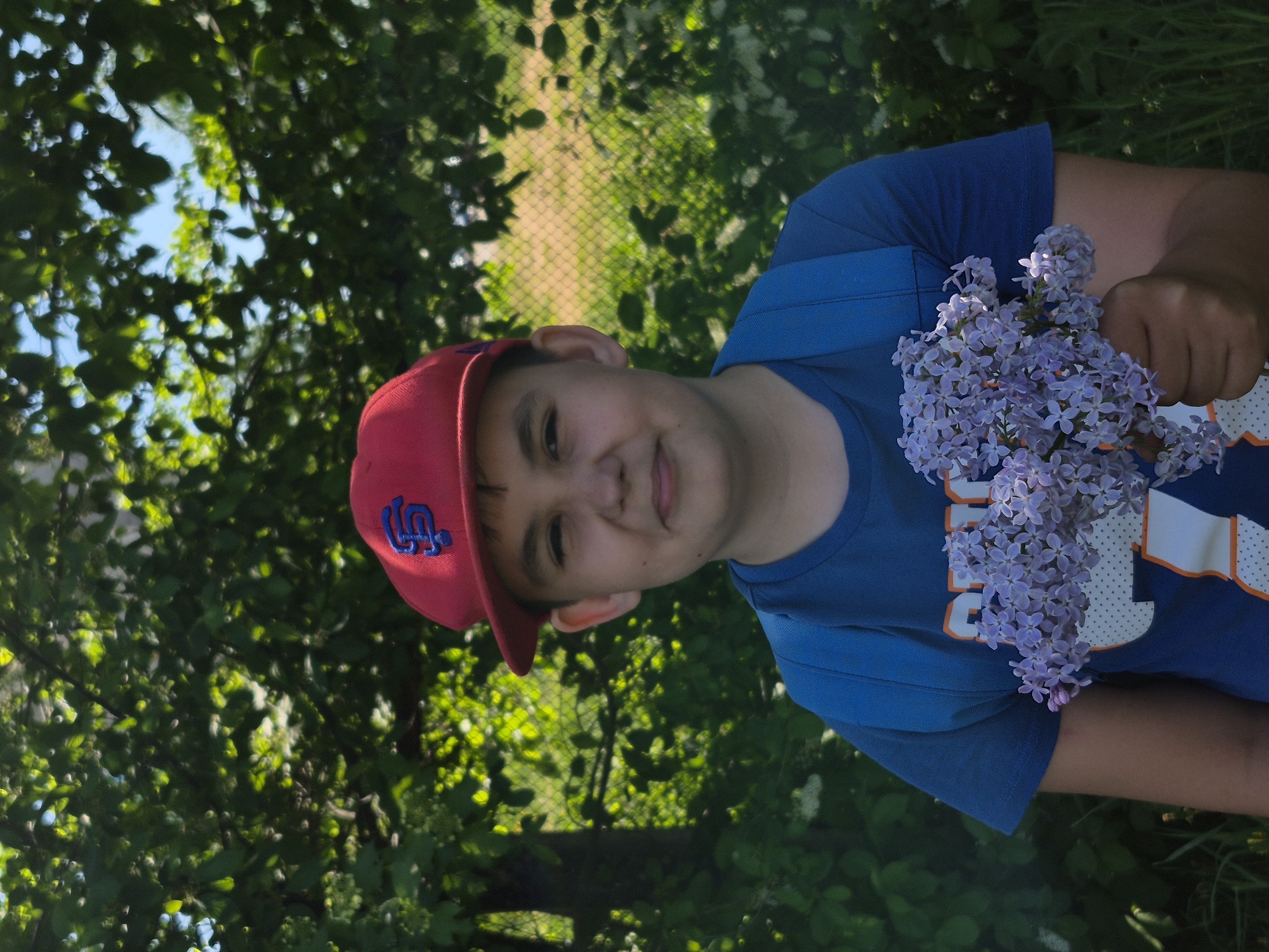 A young person is holding a bouquet of lilac flowers. The person is wearing a red baseball cap with a blue logo, a blue t-shirt, and has a backpack on. The background features green foliage, possibly trees, and a glimpse of what appears to be a fence. The location is Chaikovsky, Russia.