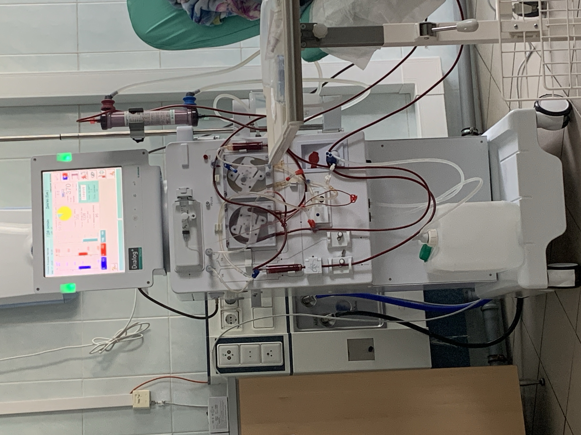 The image depicts a hemodialysis machine, possibly in a medical facility. The machine has a screen displaying various parameters, with the word "Dialog+" visible. Red and clear tubes connect to different parts of the machine. A filter and a white container are also visible. The machine is mounted on a wall with electrical outlets and other connections. The location is reported to be Chaikovsky, Russia, although this is not verifiable from the image itself. The environment appears to be a clinical setting, based on the tiled walls and the nature of the equipment. No people are visible in the image.