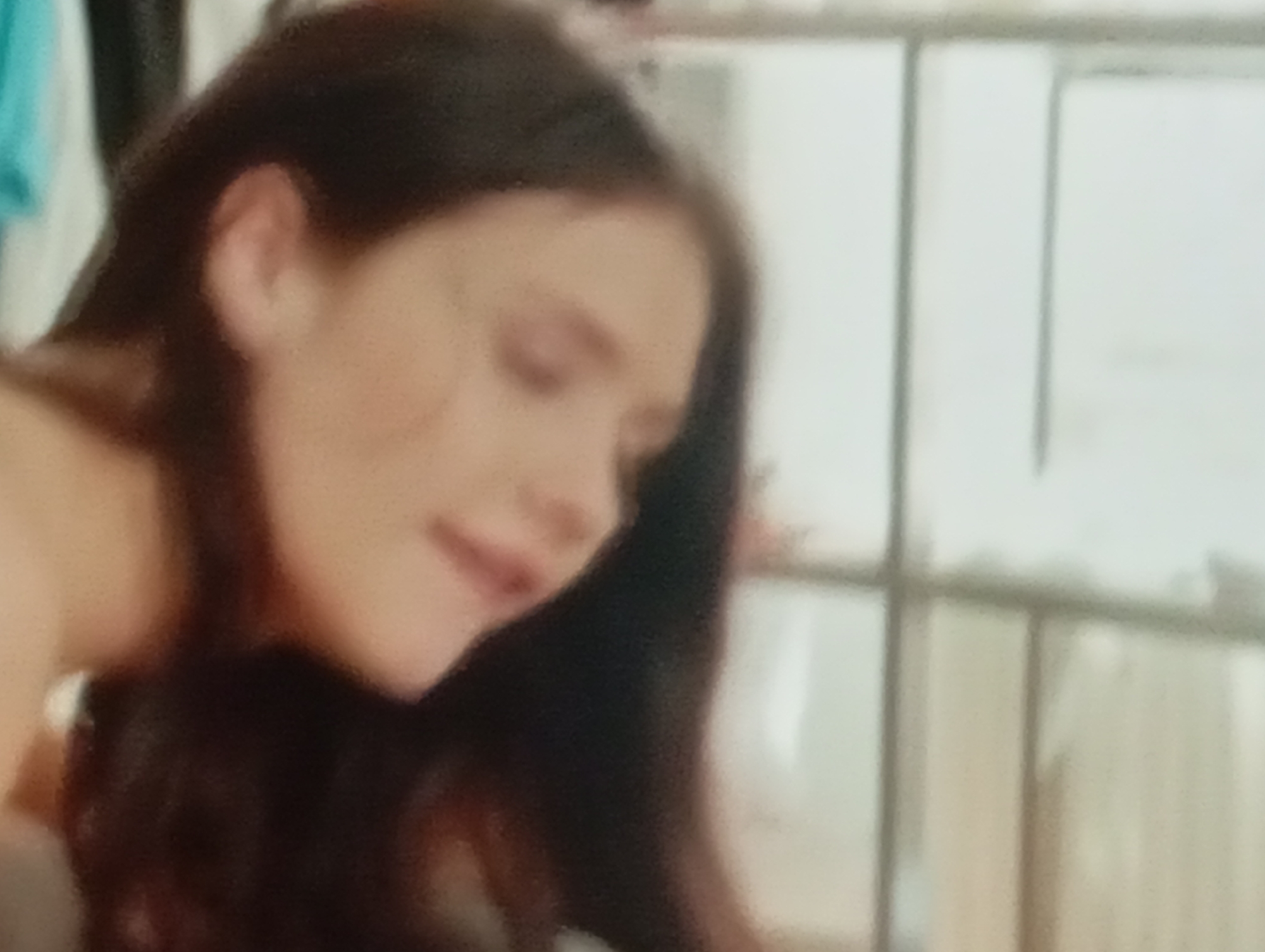 The image is a close-up, blurry shot of a person's face, with a focus on the left side of their head and neck. The person appears to have long, dark hair. The background is out of focus, but suggests an indoor setting with some sort of shelving or furniture behind. There are no visible landmarks or details that would associate the image with Sofiivska Borshchahivka, Ukraine.
