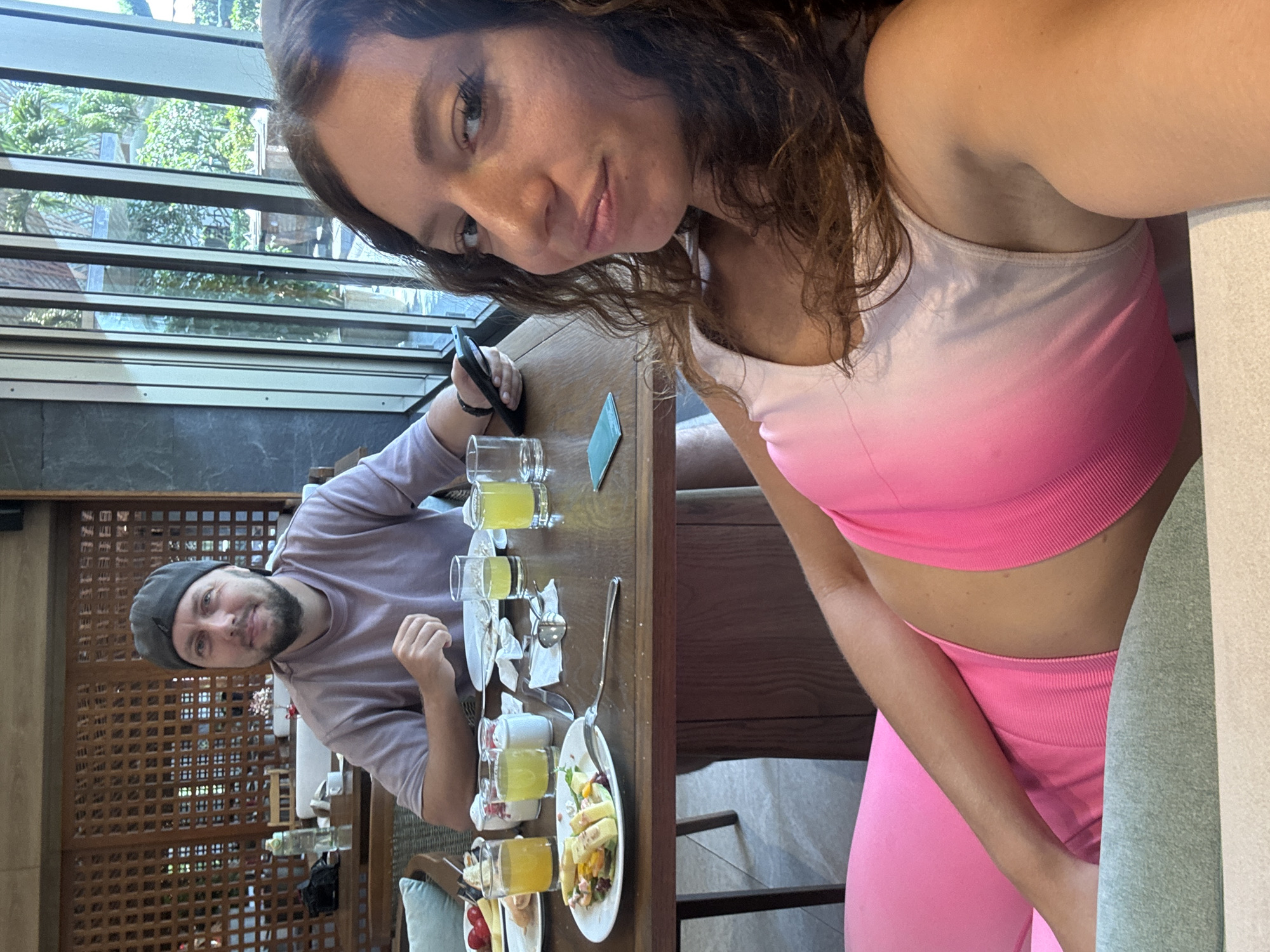 Two people are at a table in a restaurant. The woman, with brown hair, is wearing a pink sports bra and leggings and appears to be taking a selfie. The man is wearing a gray beanie and lavender-colored shirt. They are seated at a table with drinks and food, including a plate of salad. The restaurant has a window or skylight above. The location is likely in Đà Nẵng, Vietnam.