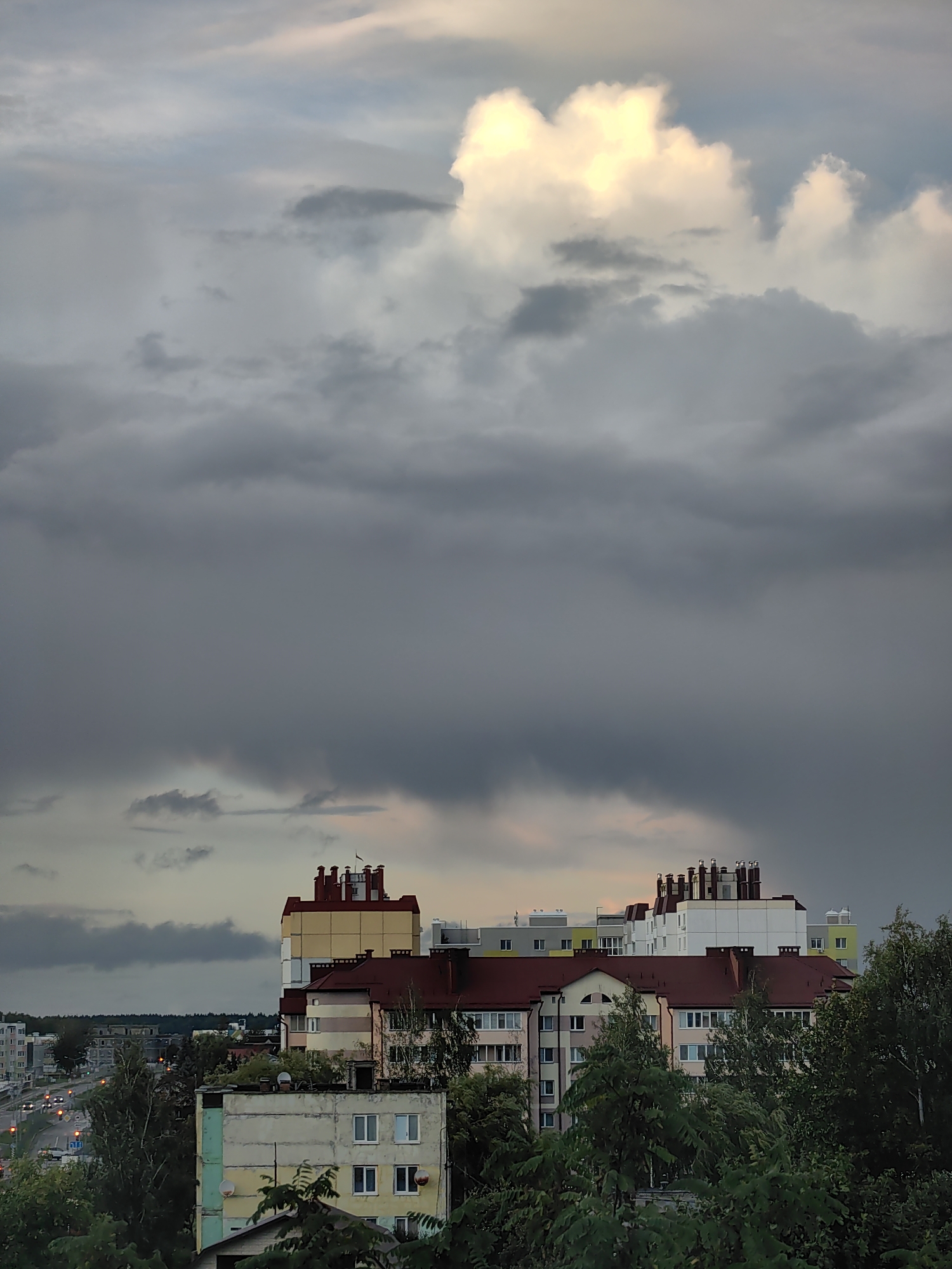 An exterior, eye-level, full shot captures a dense, overcast sky above a cluster of multistory buildings and trees in Fanipal, Belarus. The sky is dominated by dark, heavy clouds that obscure most of the sun, though a bright patch of lighter clouds and a sliver of sunlit sky are visible towards the top center. Below the clouds, a line of residential buildings with red tiled roofs and colorful facades is visible. The buildings vary in height and design, with some featuring balconies and numerous windows. In the foreground, dense green foliage from trees partially obscures the lower portions of the buildings. To the left, a street with traffic and streetlights can be seen, indicating an urban environment. The overall impression is one of impending rain or a recent storm. There are no people or animals visible in the frame.