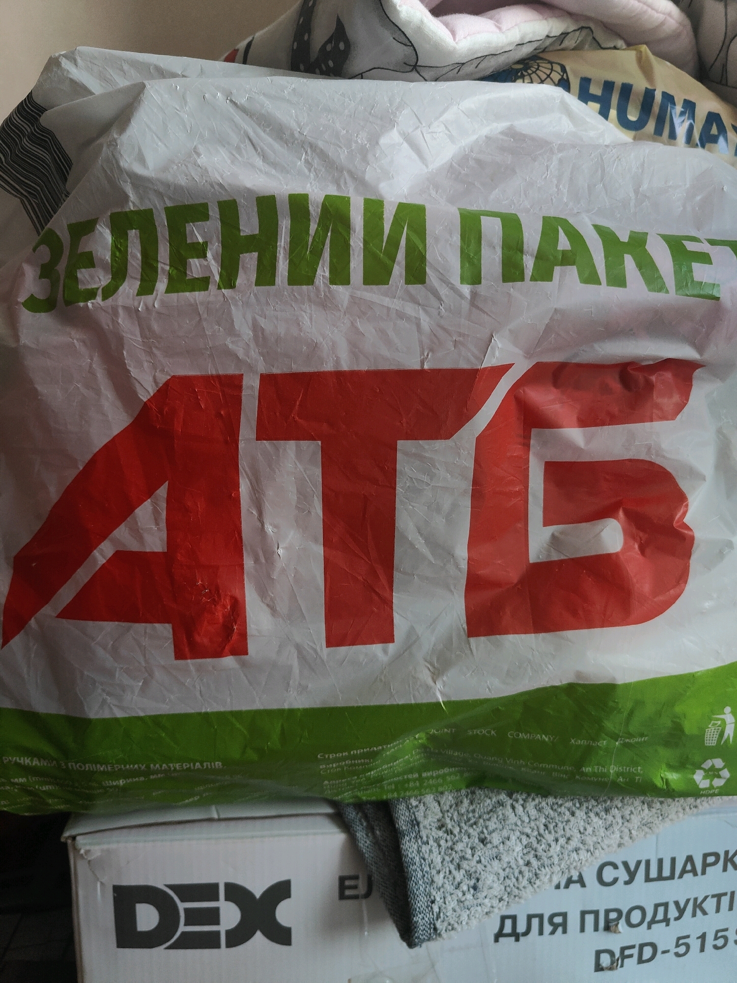 The image shows a white plastic bag with large red and green lettering. The red lettering appears to say "ATG". The green lettering says "ЗЕЛЕНИЙ ПАКЕТ" which translates to "Green Package". Below this, in smaller text, are details about the bag's materials and manufacturer, including "JOINT STOCK COMPANY" and an address in Vietnam. There is also an image of a person recycling and a "HDPE" symbol. The bag is positioned on top of a white cardboard box. The box has the "DEX" logo in large black letters and some Ukrainian text. The background is blurred and appears to be some sort of fabric. The scene is likely indoors, given the lighting. There are no people or actions depicted in the image. The location in Chornomorsk, Ukraine, is not directly indicated by any visual cues within the provided image.
