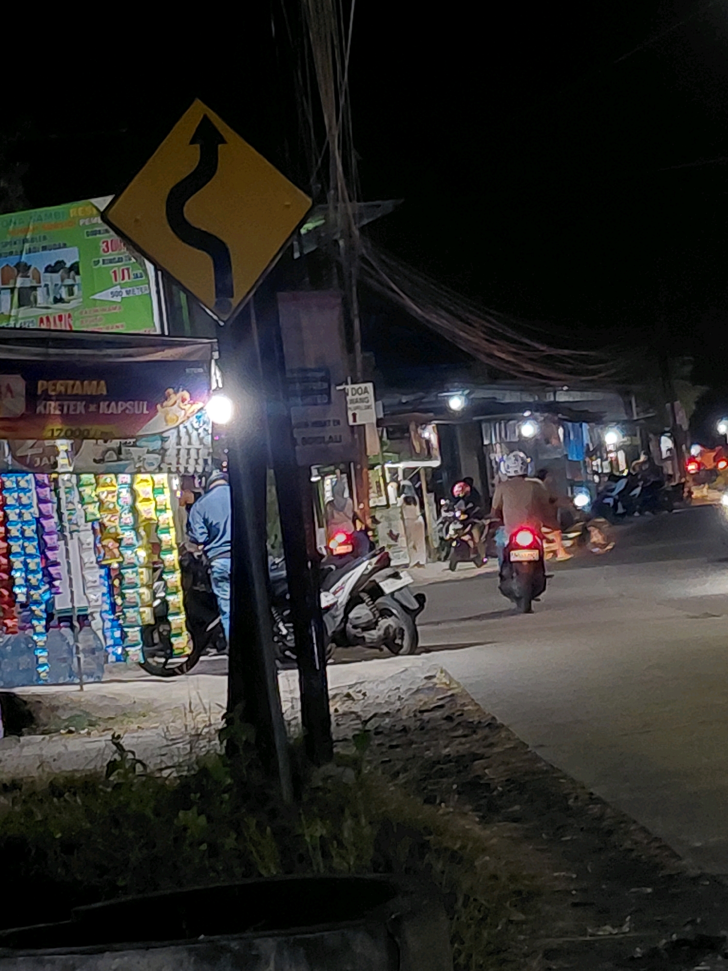 In Babadan, Indonesia, a nighttime street scene unfolds with a yellow diamond-shaped traffic sign featuring a black serpentine arrow pointing upwards and to the right, indicating a winding road ahead. To the left of the sign, a store displays various products, including cigarettes and snacks, illuminated by artificial lights. A man in a blue shirt and jeans stands in front of the store, facing away from the camera. A motorbike with a rider is parked nearby. Further down the street, several other motorbikes and their riders are visible, some in motion and others stationary. The street is paved, and the surrounding environment appears to be a commercial area with small shops and overhead wires. The lighting conditions are dim, with artificial lights creating strong contrasts and casting shadows.