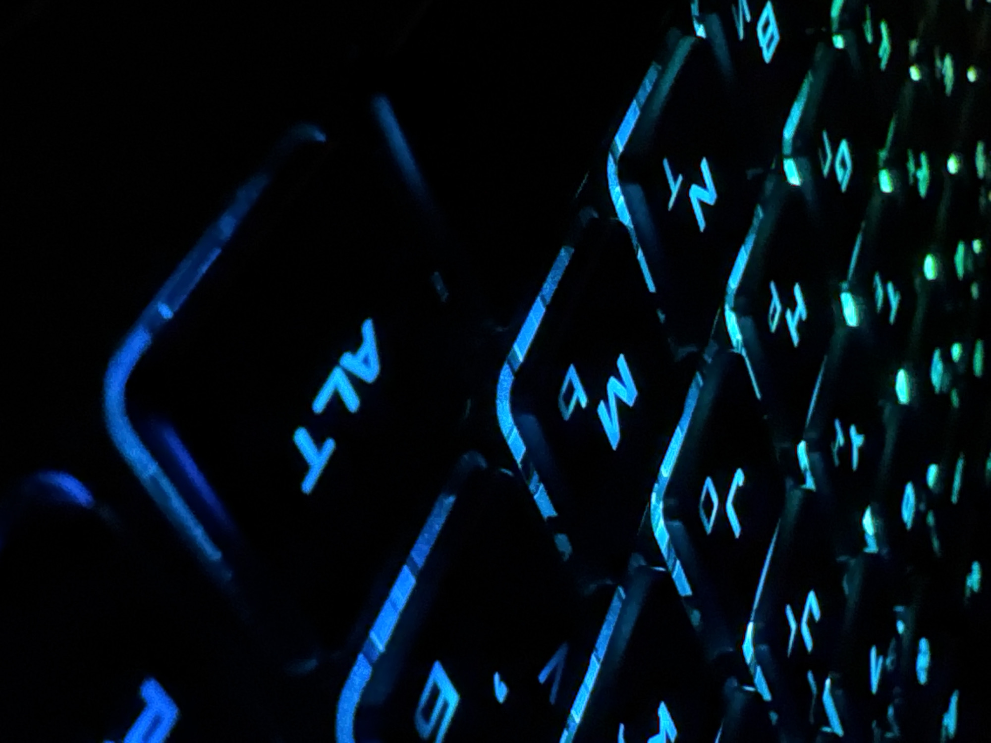 The image is a close-up, low-light photograph of a keyboard with blue and green backlighting. The keys are black with illuminated symbols and letters. Visible keys include "N", "T", "M", "b", "ALT", and parts of other keys with partially visible symbols. The lighting creates a bokeh effect, with the background being out of focus and dark. The scene appears to be an indoor setting, likely a desk with a computer. There are no people or animals present in the image, and no specific location in a city is identifiable. The focus is on the illuminated keys and the texture of the keyboard.