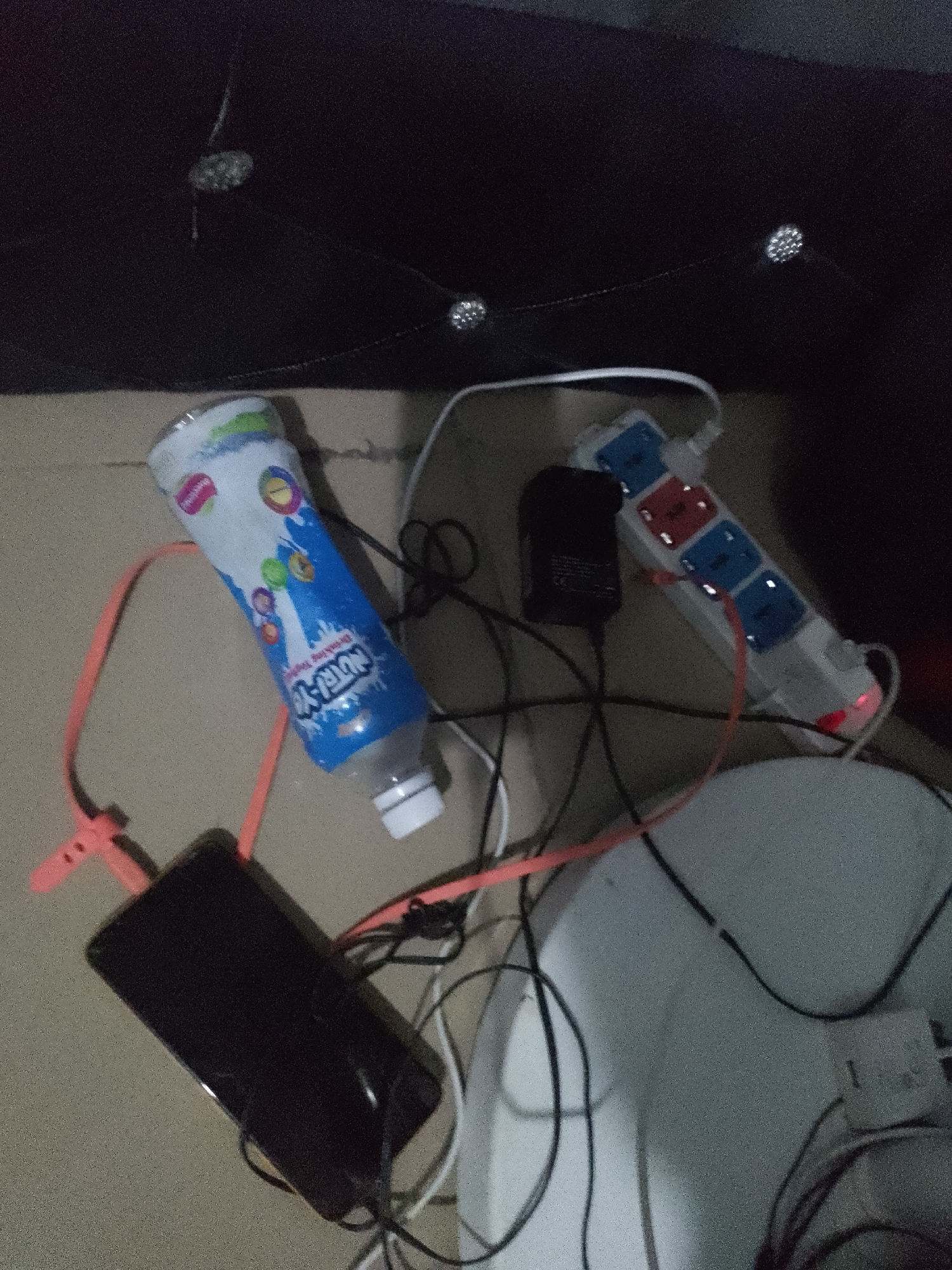 In a dimly lit indoor environment, possibly a bedroom, several electronic devices and a beverage bottle are scattered across a textured surface. A white power strip with multiple outlets, some red and some blue, is connected to various cords. A black power adapter is plugged into one of the outlets. A smartphone, lying face down, is connected to a charging cable. To the left, a blue and white plastic bottle with colorful graphics, identified as "Nutri-Yo," is placed on its side. The background is a dark, textured surface, possibly upholstery, with decorative circular embellishments. The overall scene suggests a messy charging station or workspace.