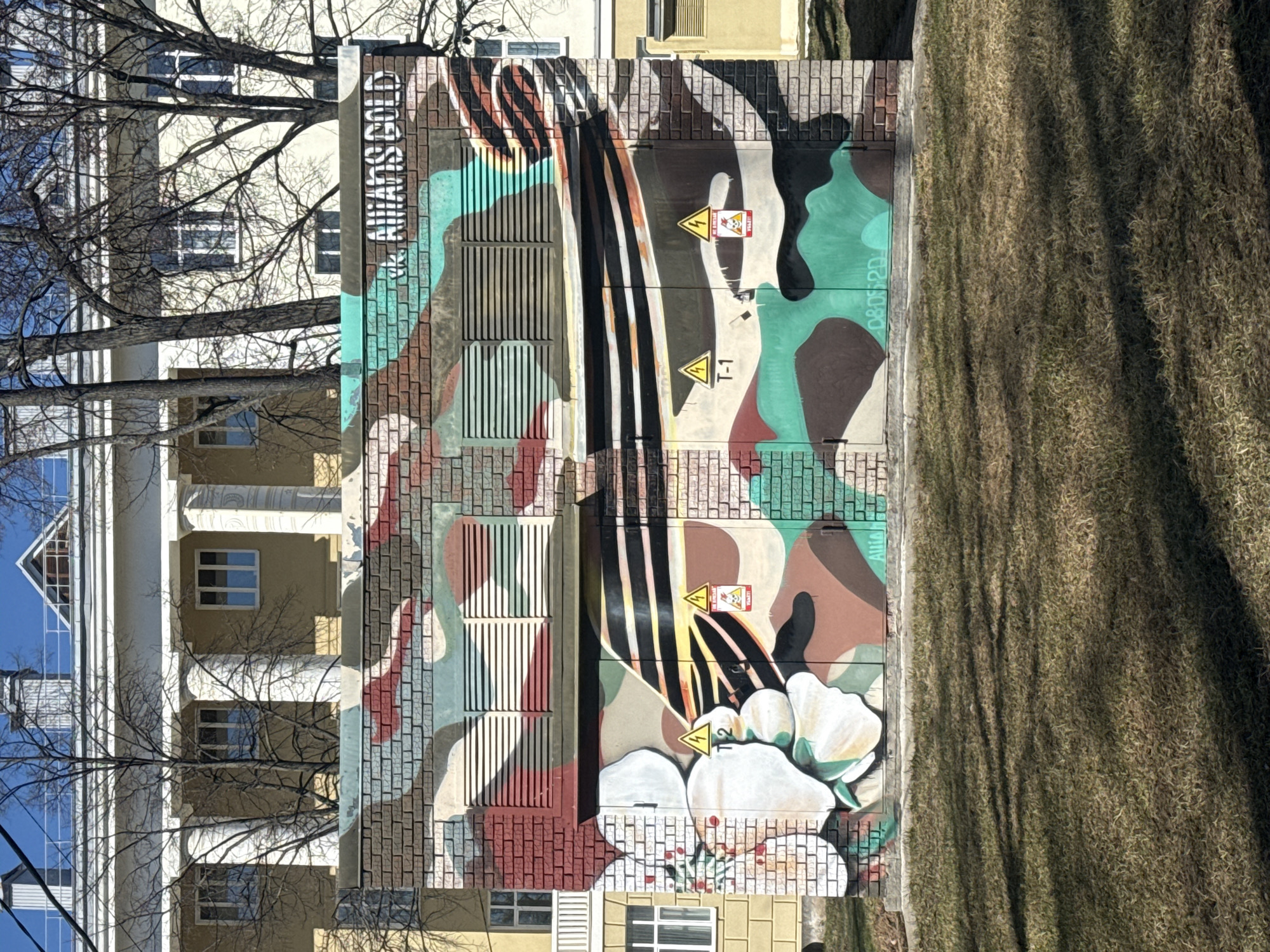 An eye-level outdoor shot shows a utility box painted with a graffiti-style mural in Votkinsk, Russia. The mural incorporates camouflage colors like brown, green, tan, and black, along with stripes, flowers, and yellow "Danger" signs with a lightning bolt symbol and text. The phrase "ALWAYS GOLD" is also painted on the box.
To the right of the box, a dry grass field extends. To the left, a multi-story building with white columns and beige walls can be seen. Bare trees frame the building and the blue sky is visible above.